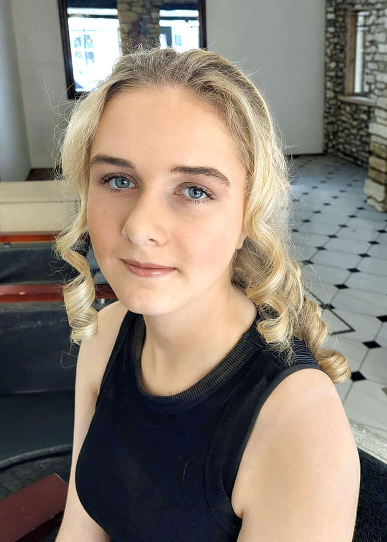 Young woman with curly blonde hair and blue eyes wearing a black sleeveless top, seated indoors with a tiled floor and stone wall in the background.
