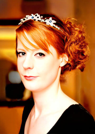 Portrait of a woman with red hair styled in a low curly bun, wearing a black top and a white floral headband.
