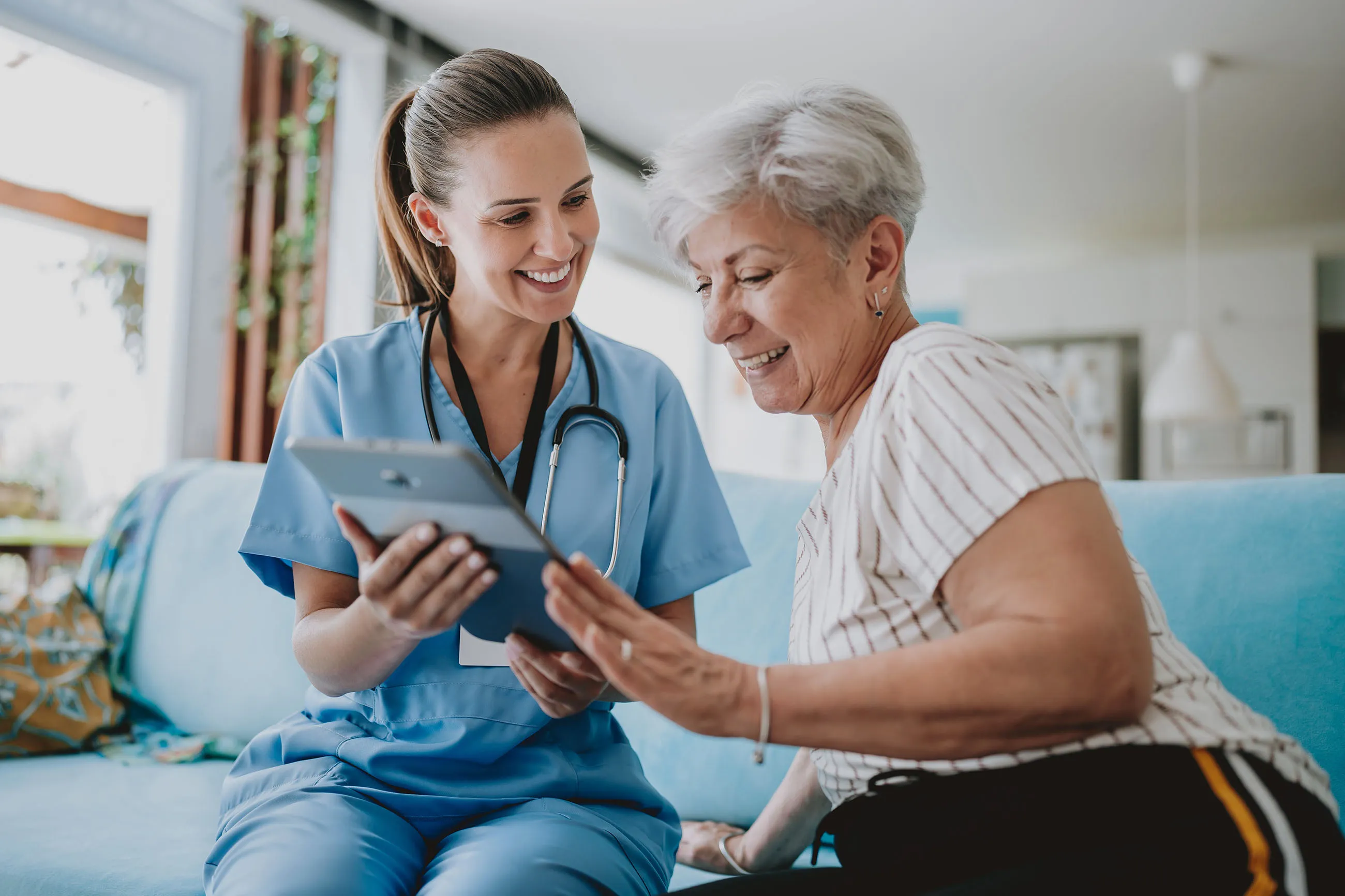 Healthcare professional in blue scrubs showing a tablet to a smiling elderly woman on a couch in a bright living room.