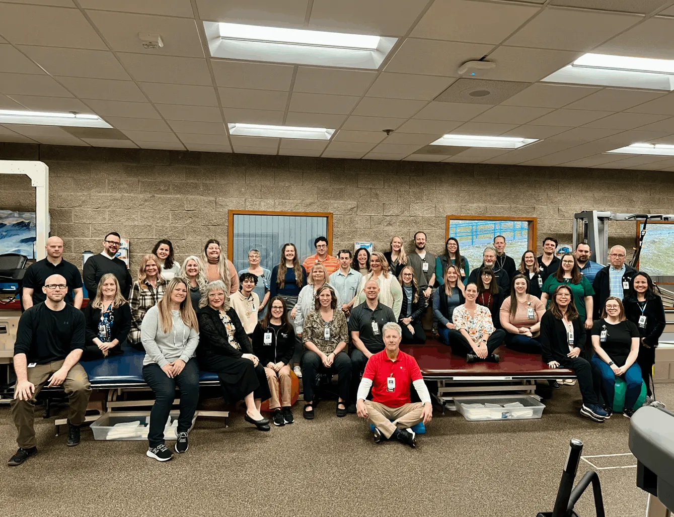 Group photo of around 40 people in a physical therapy or rehabilitation gym, some sitting on therapy tables and others standing against a brick wall with two windows.
