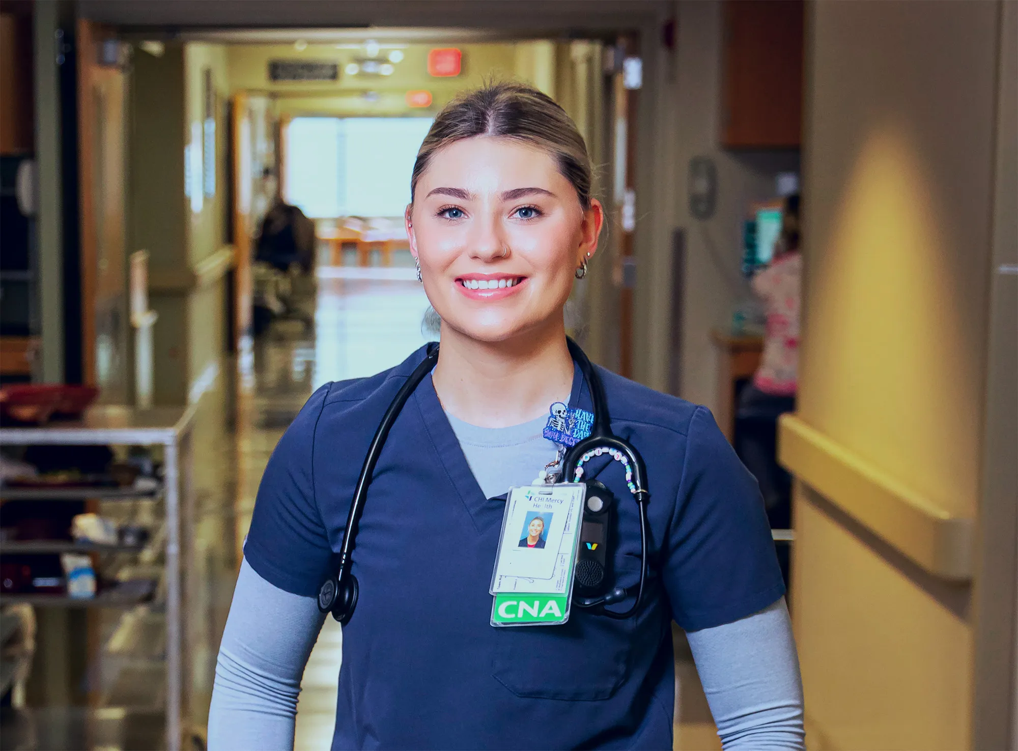 Smiling female healthcare worker in navy scrubs and stethoscope standing in a hospital hallway.