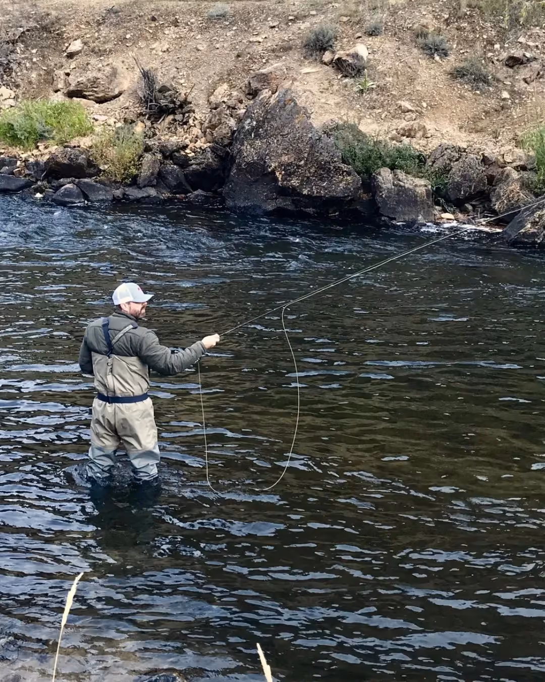 Paul Morrison fly fishing while standing waist-deep in a river with rocky bank and sparse vegetation.