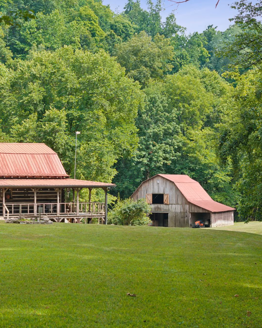 Wooden house and barn with red roofs surrounded by lush green trees and grass.