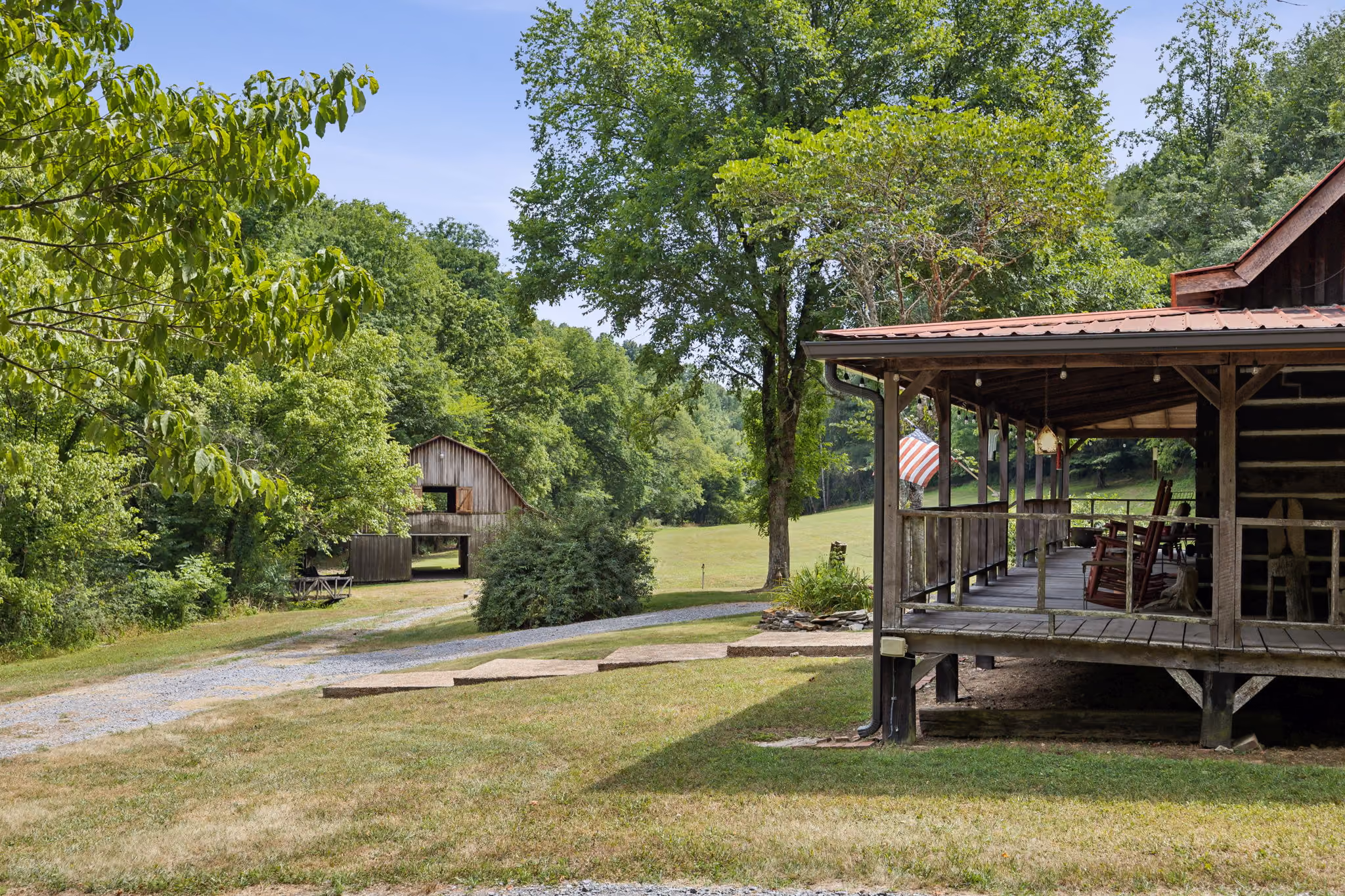 Wooden cabin porch with rocking chairs and American flag overlooking grassy yard and barn.