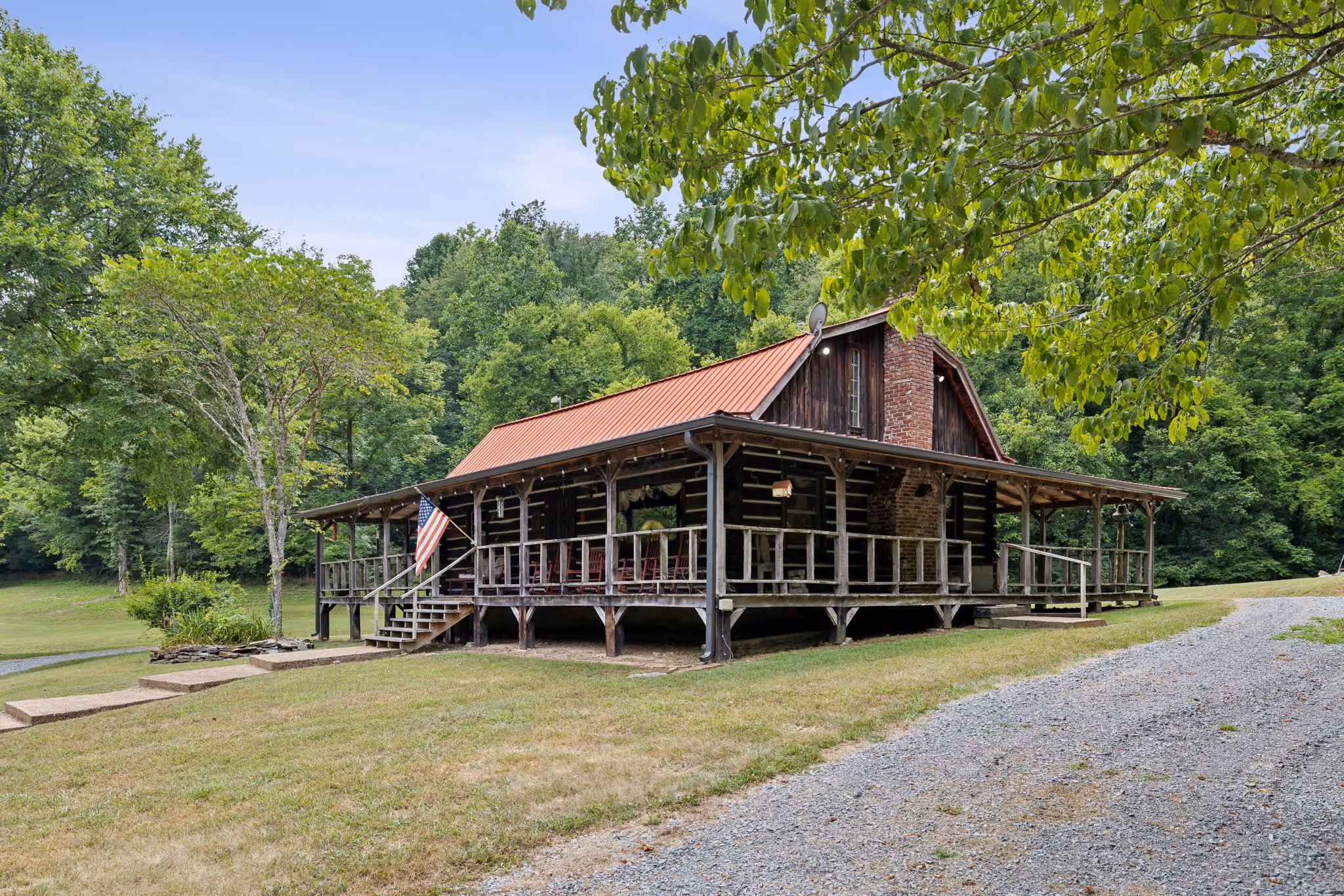 Rustic log cabin with wraparound porch, red metal roof, and American flag in green wooded area.