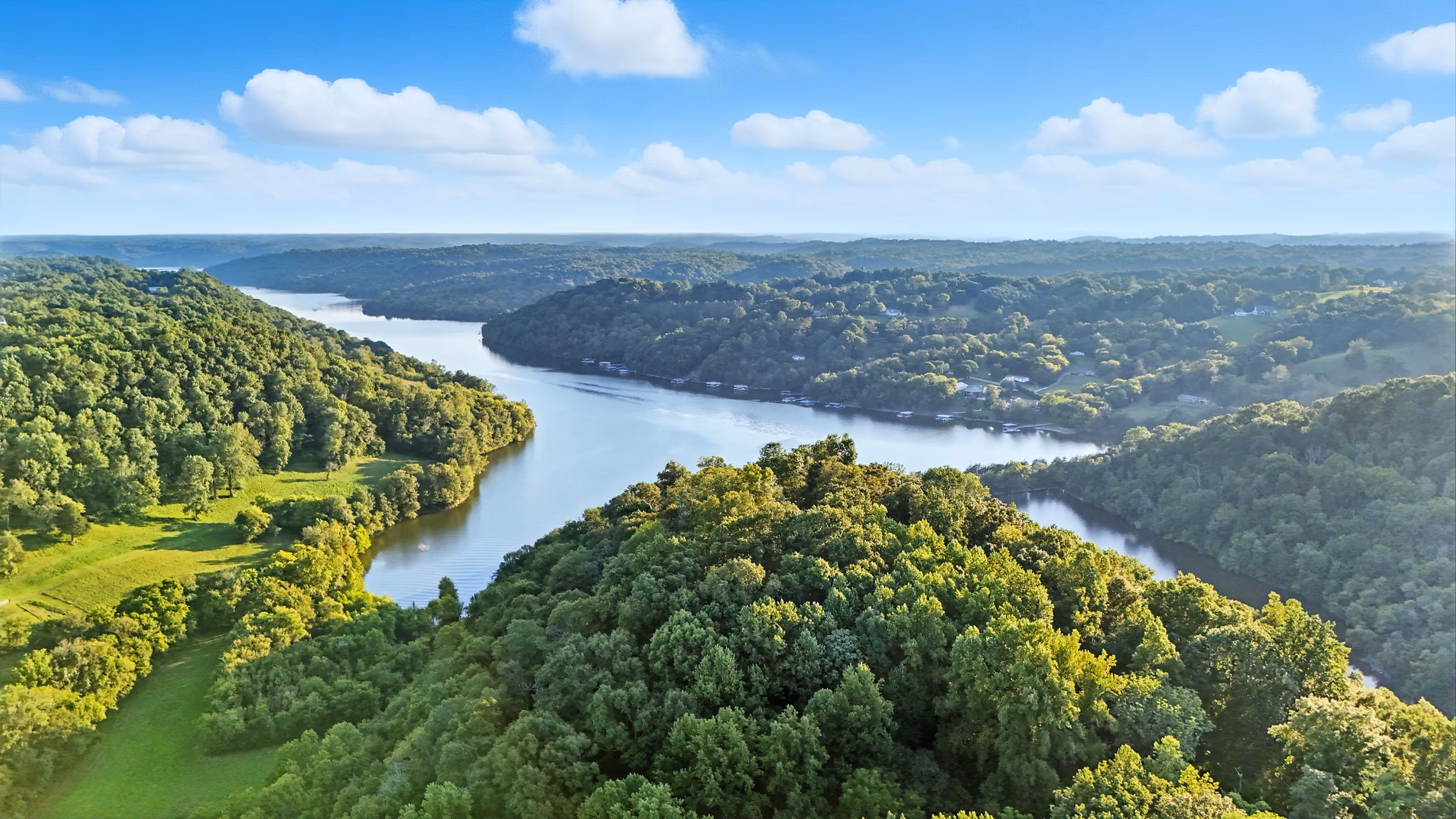 Aerial view of a winding river surrounded by dense green forests and hills under a blue sky.
