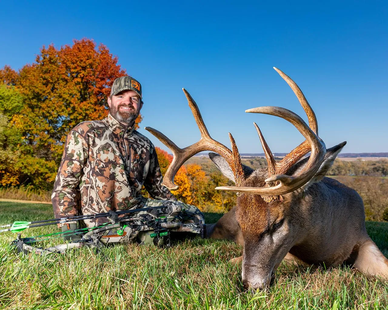 Paul Morrison in camo gear sits next to large deer with big antlers on grassy field in fall.