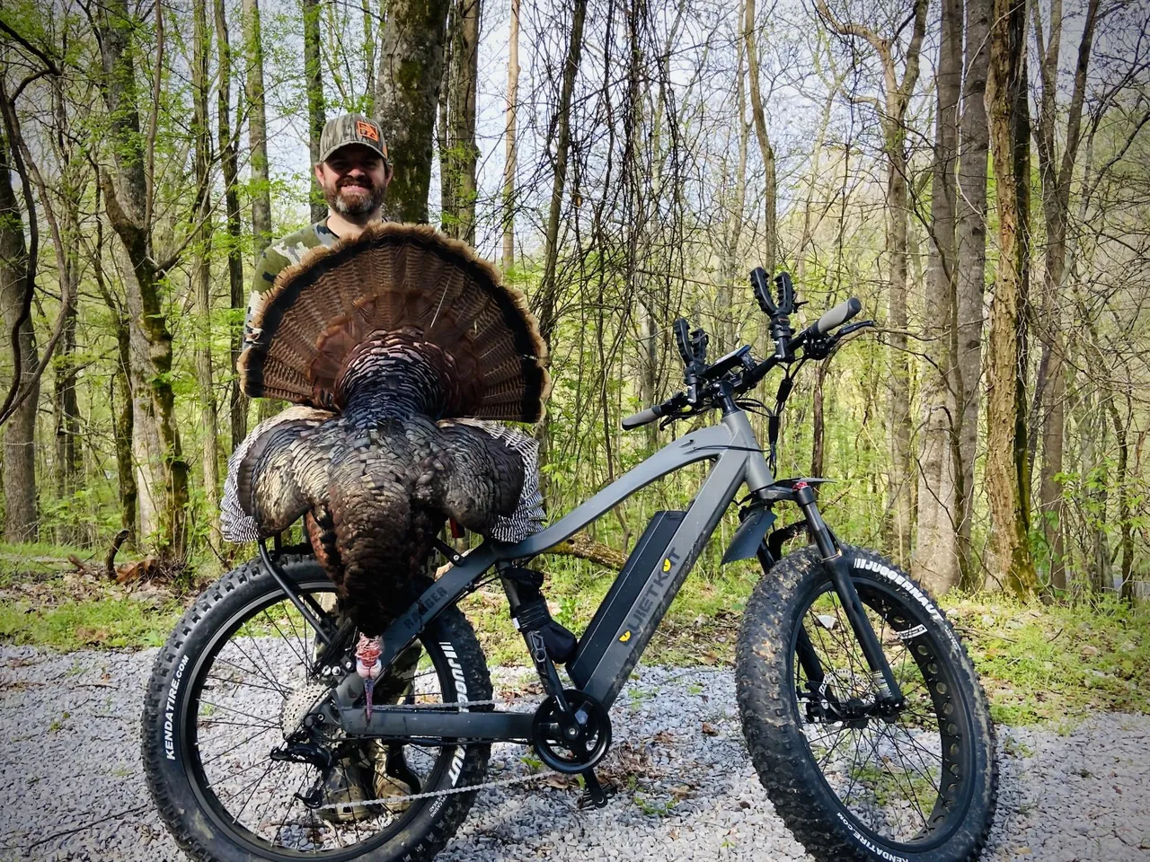 Paul Morrison wearing camouflage holding a large turkey behind an electric fat-tire bike on gravel in the woods.