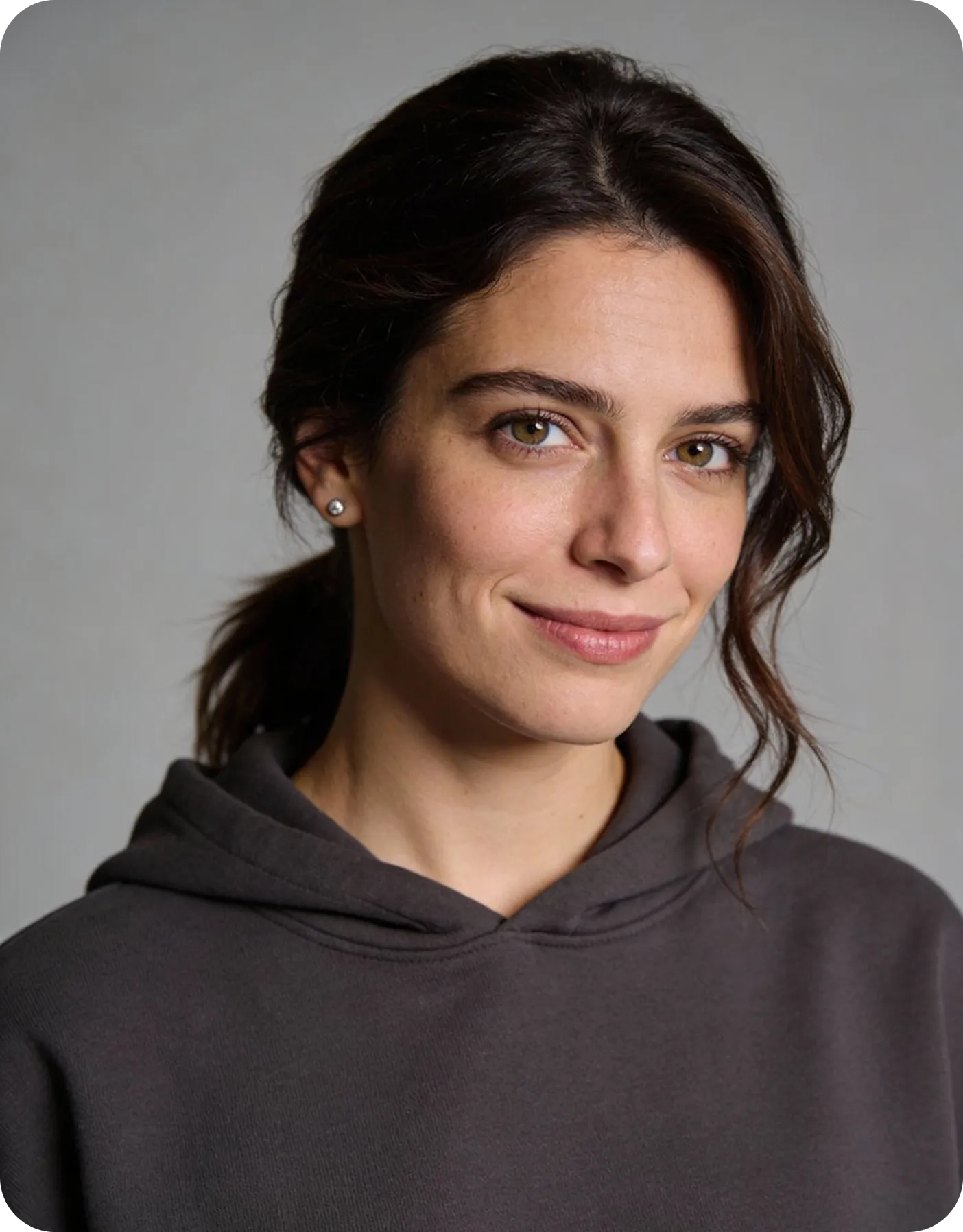 Young woman with brown hair in a ponytail, wearing a dark hoodie and smiling gently against a plain gray background.