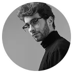 Black and white photo of a man with glasses and wavy hair wearing a black turtleneck looking at the camera.