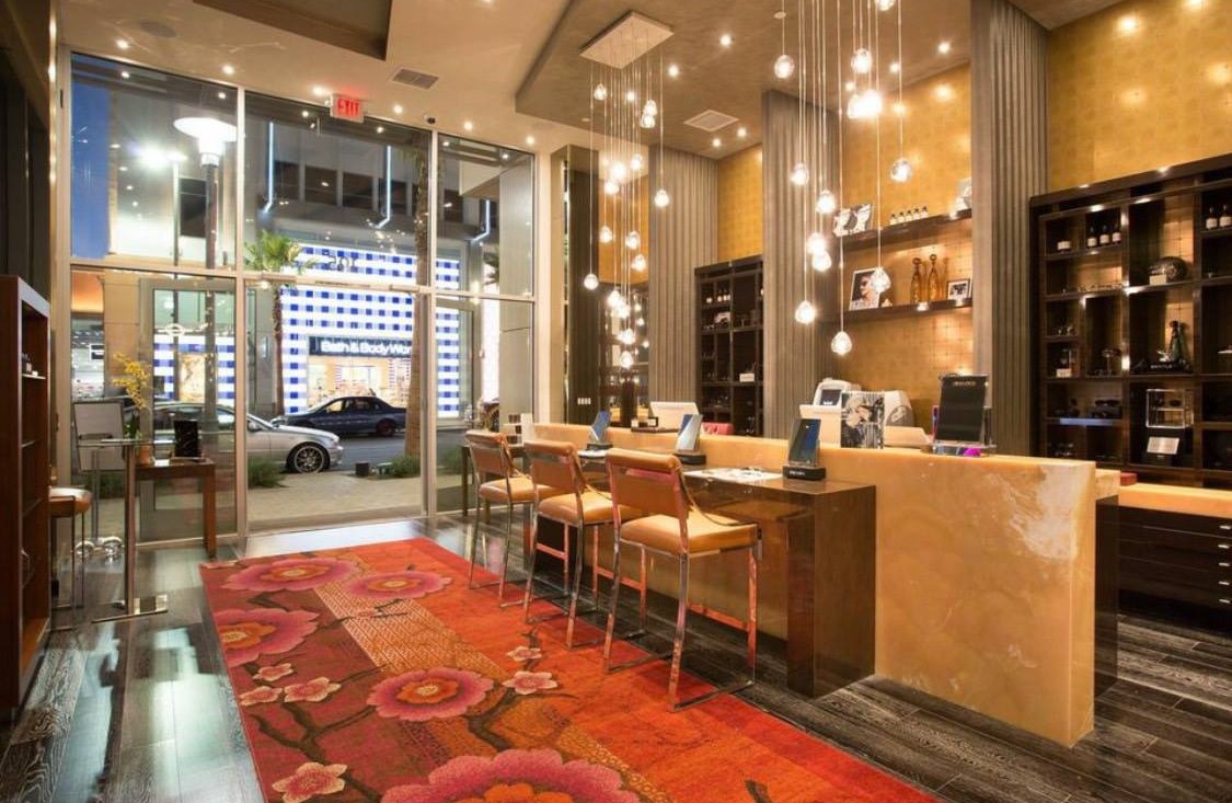 Luxurious optical boutique interior with hanging pendant lights, wooden chairs, marble counter, and a red floral carpet near the glass entrance.