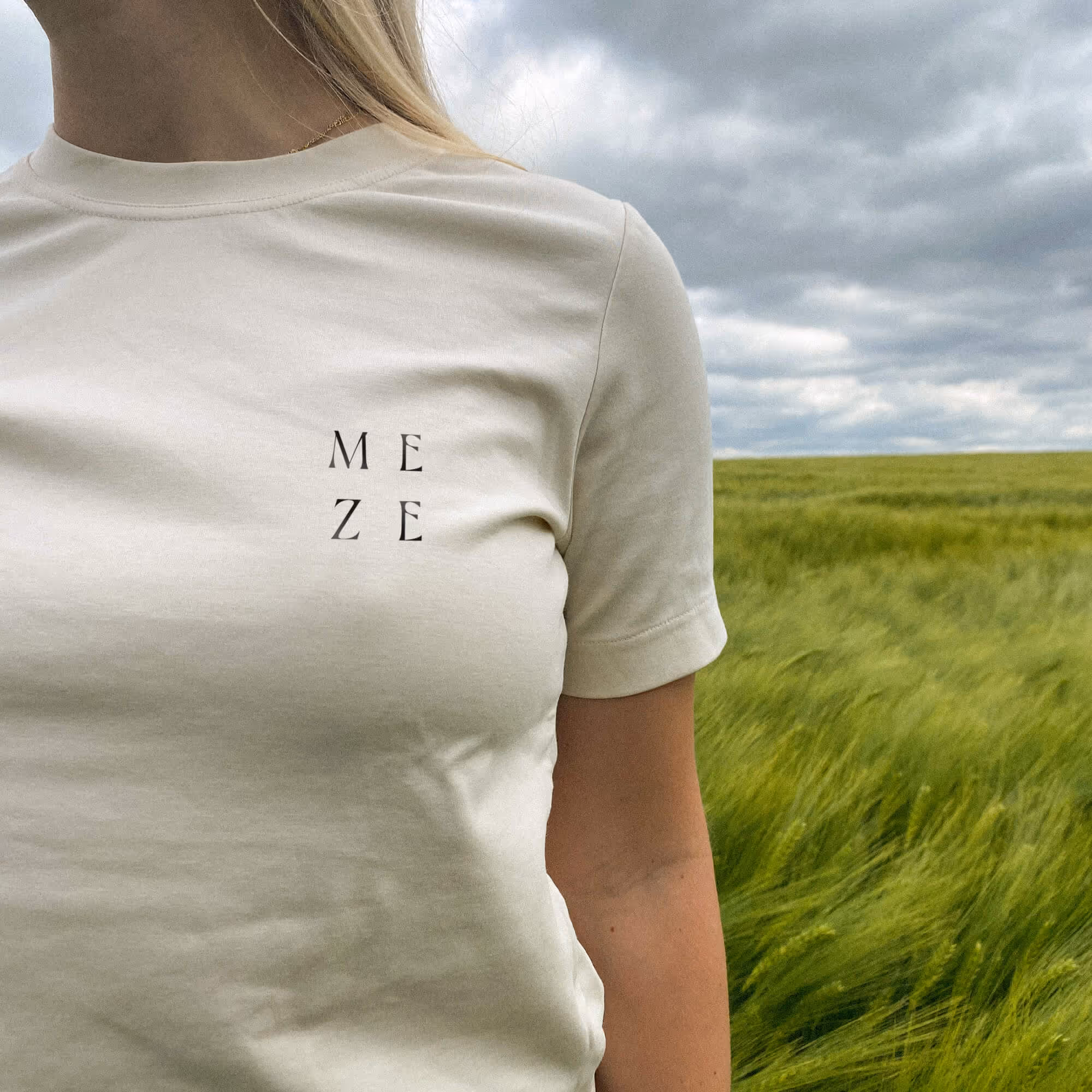 Žena v světlém tričku s nápisem MEZE stojí v poli s zelenou travou pod zataženou oblohou.