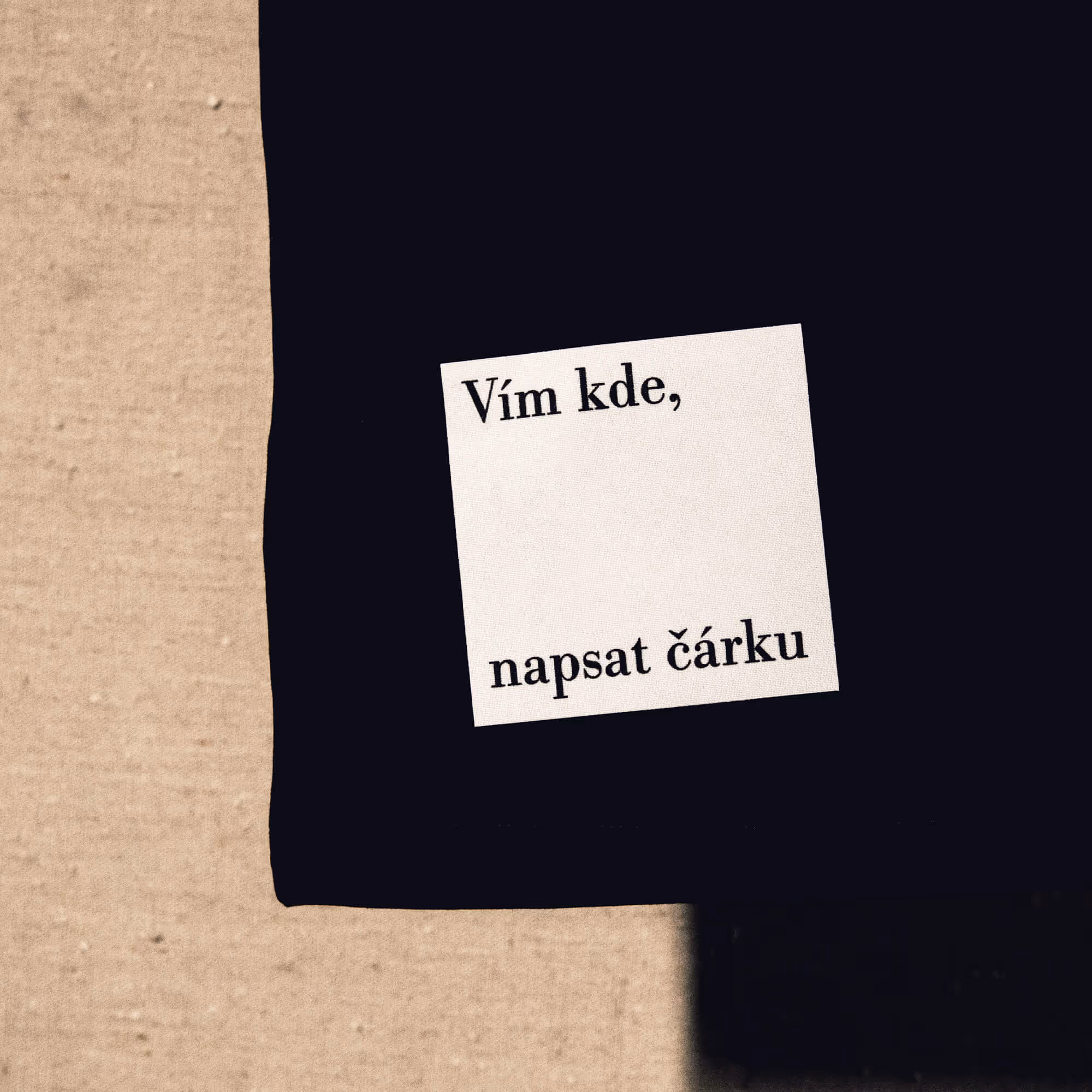 Černý kousek látky s bílou nálepkou obsahující český text „Vím kde, napsat čárku.“