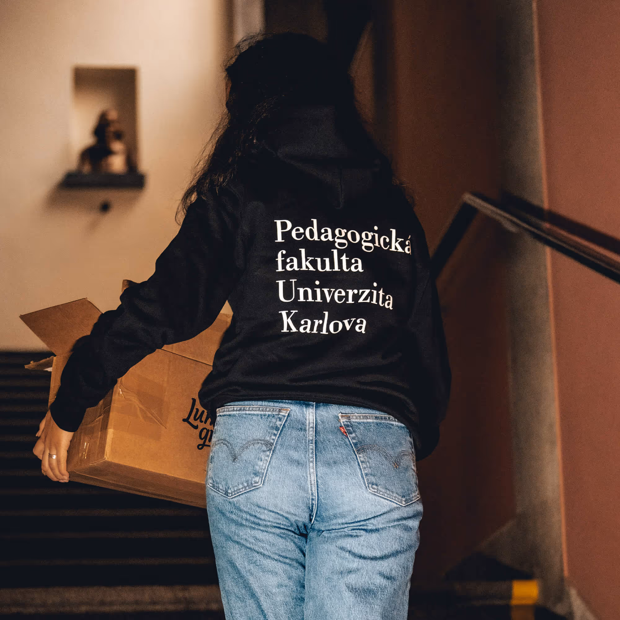 Osoba nesoucí krabici stoupá po schodech, má na sobě mikinu Pedagogická fakulta Univerzita Karlova.