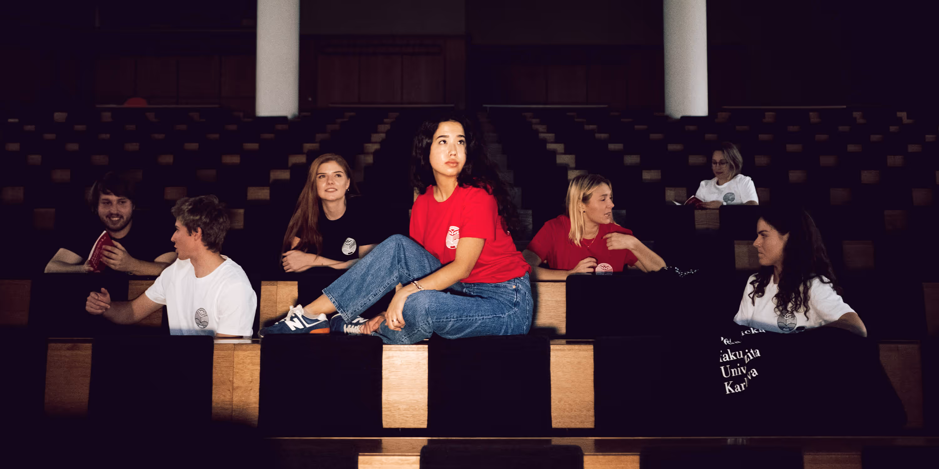 Skupina studentů sedí a komunikuje v auditoriu s dřevěnými a černými sedadly.