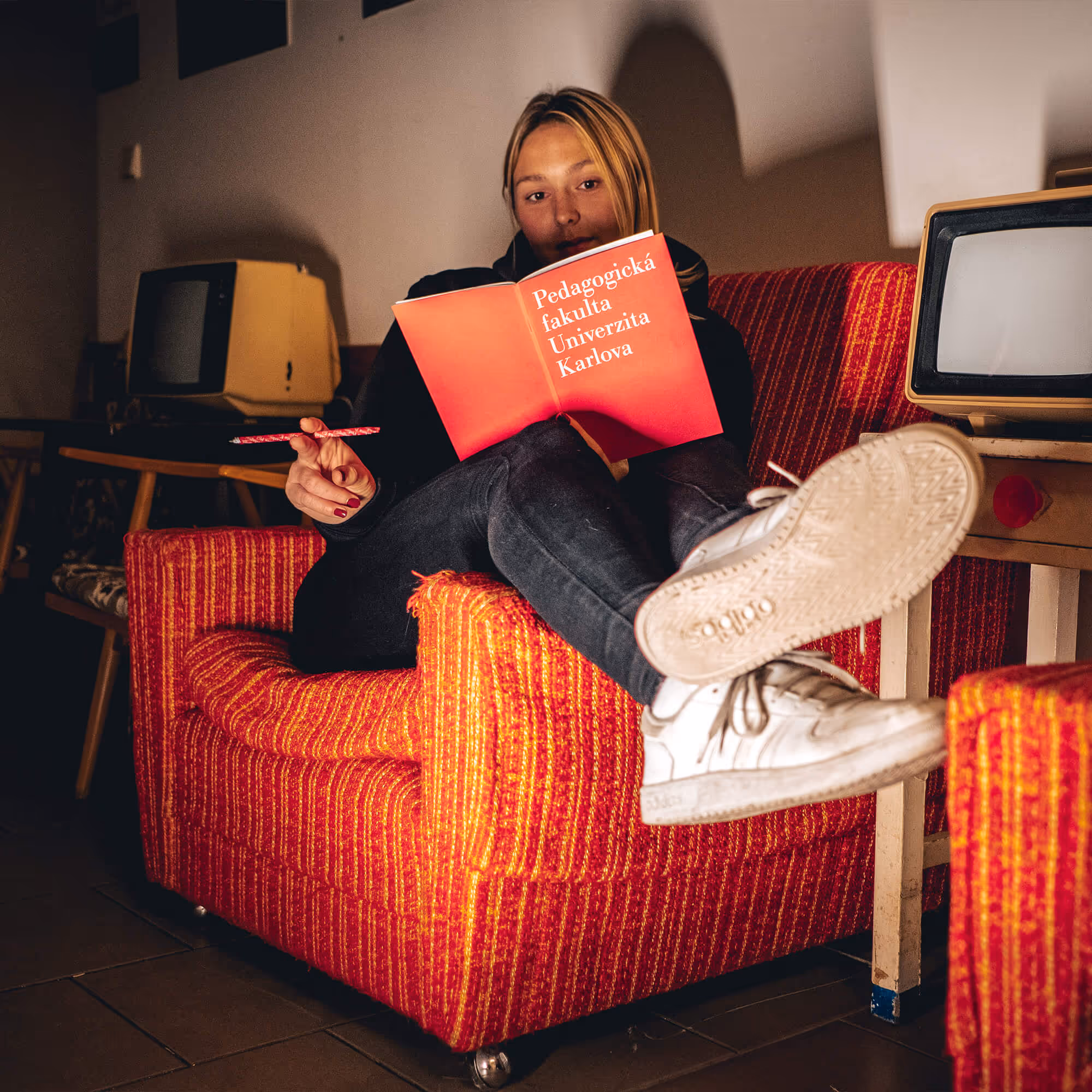 Žena sedí na červeném křesle a čte červenou knihu Pedagogická fakulta Univerzita Karlova.