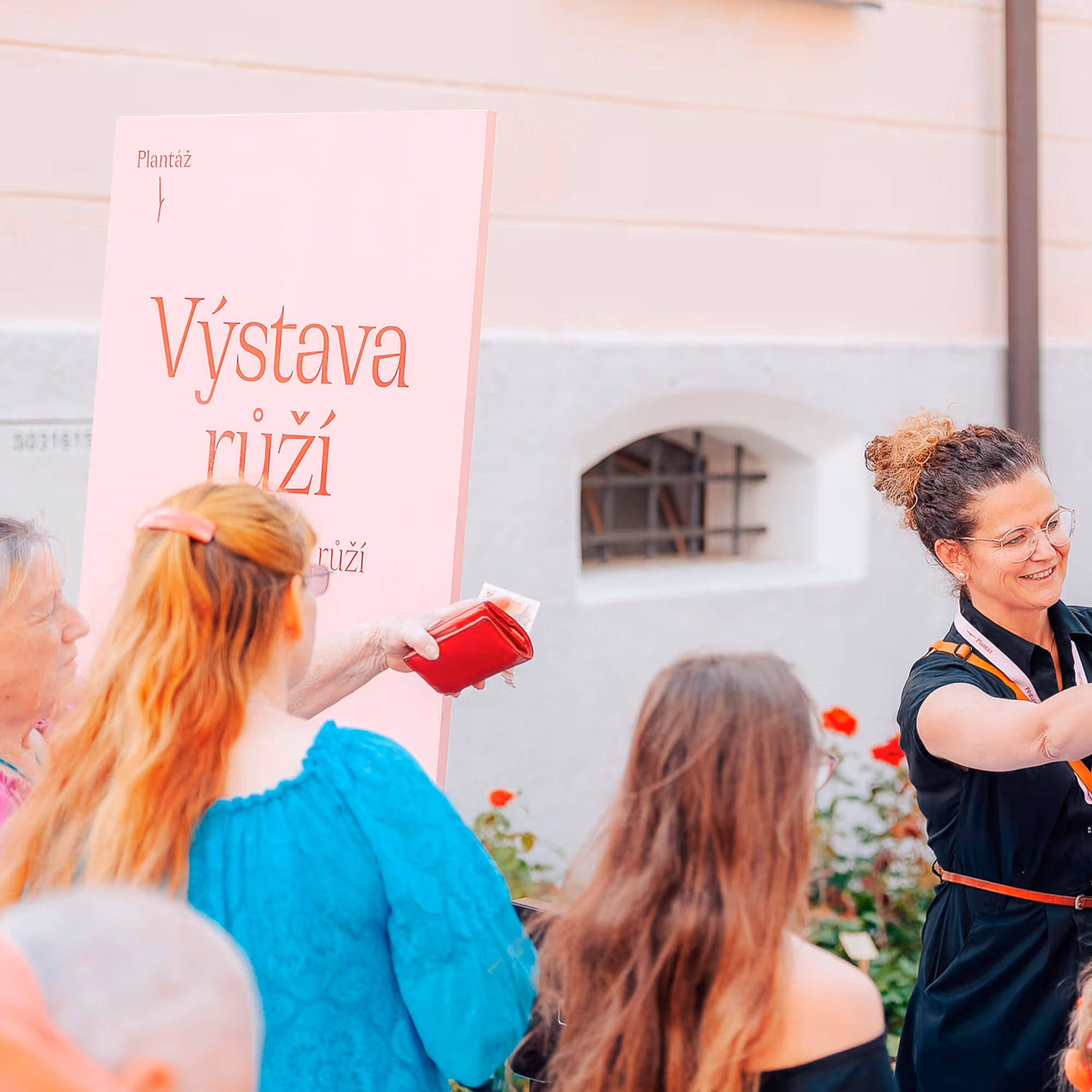 Žena s brýlemi a kudrnatými vlasy vede skupinu lidí u růžového plakátu Výstava růží.