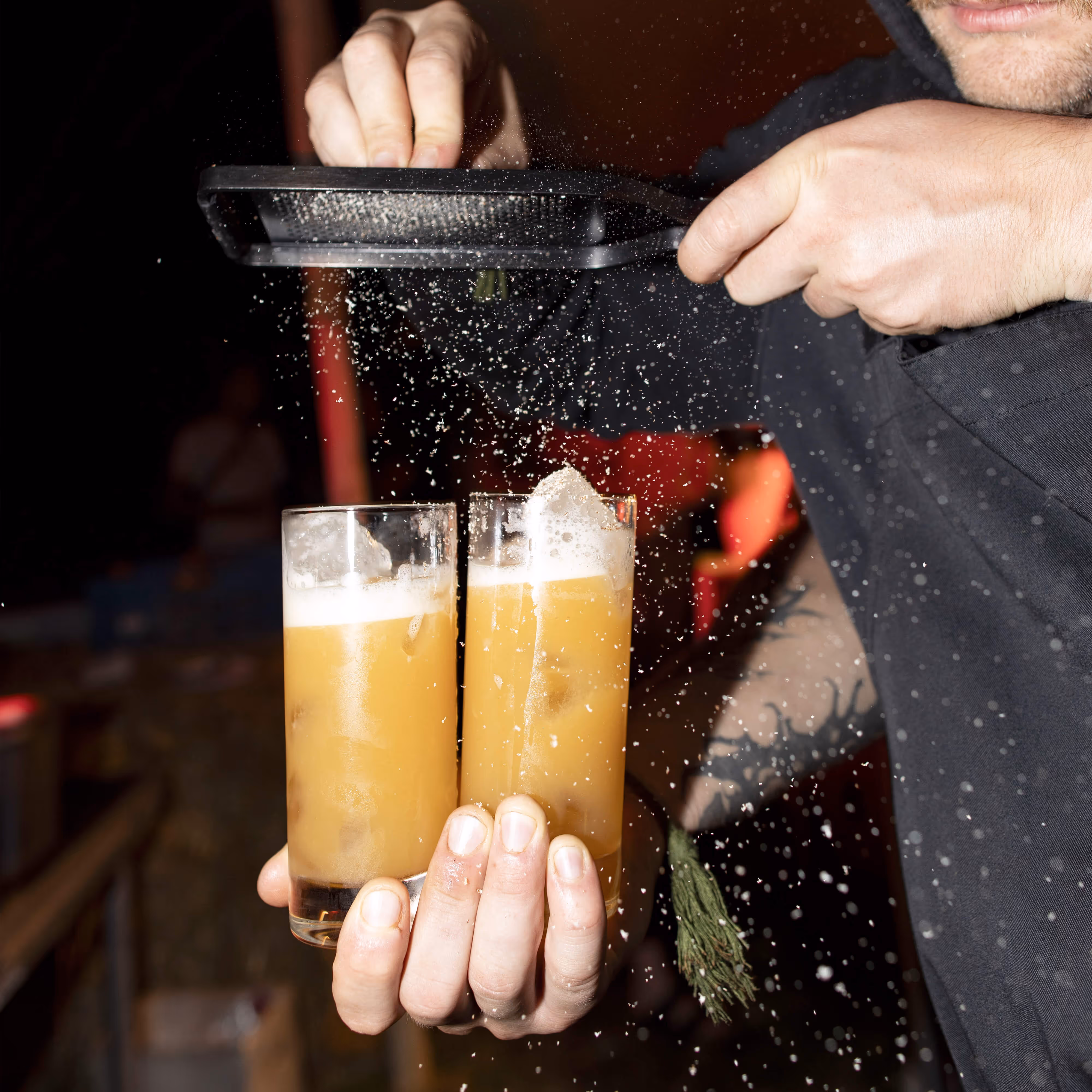 Barman posypává dva vysoké sklenice s oranžovým nápojem drceným ledem.