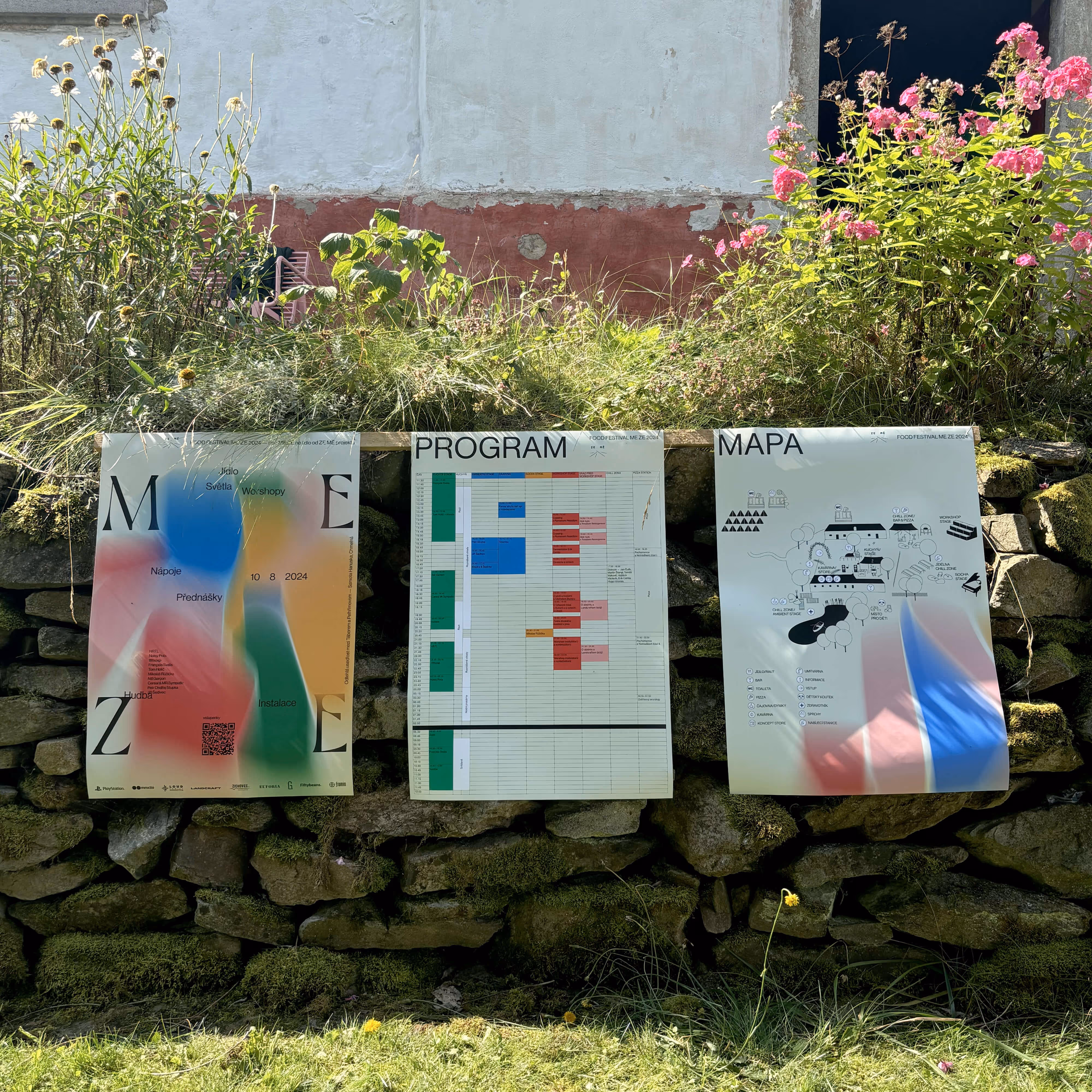 Tři plakáty festivalu se programem, mapou a uměleckou grafikou připevněné na kamenné zdi s květinami.