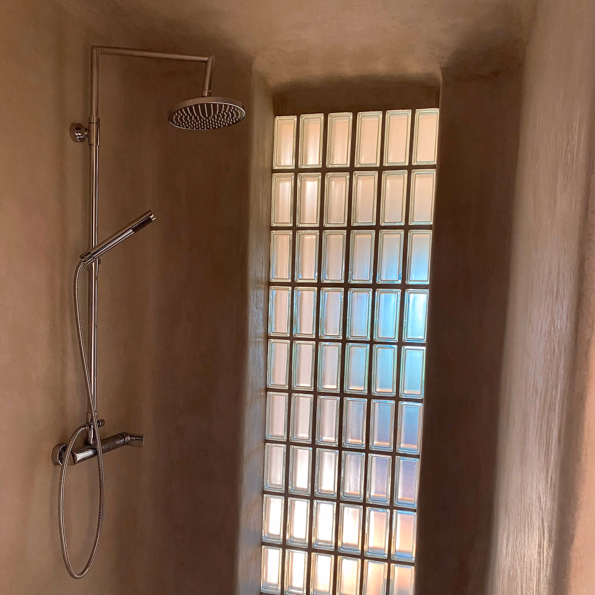 Modern shower setup with chrome rain and handheld showerheads beside a frosted glass block window.