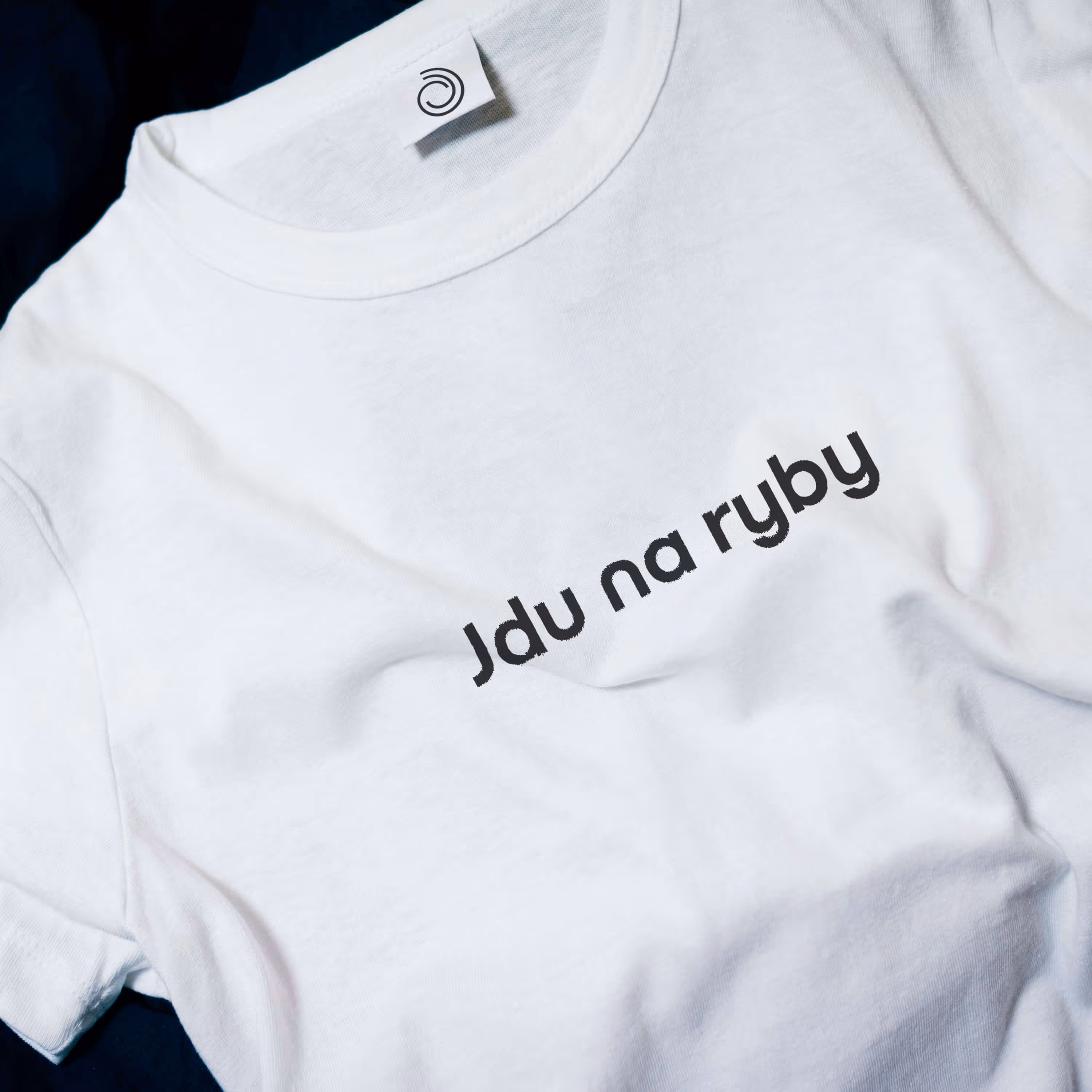 Bílé tričko s černým nápisem Jdu na ryby ležící na tmavém pozadí.