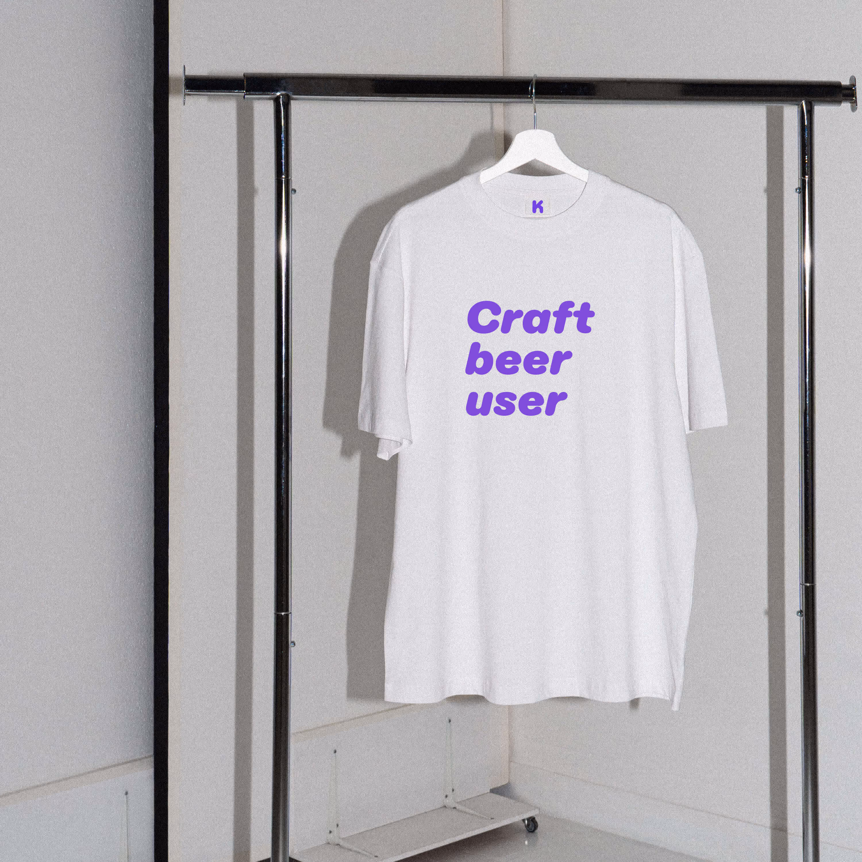 Bílé tričko na ramínku se stojanem s nápisem Craft beer user fialovou barvou.
