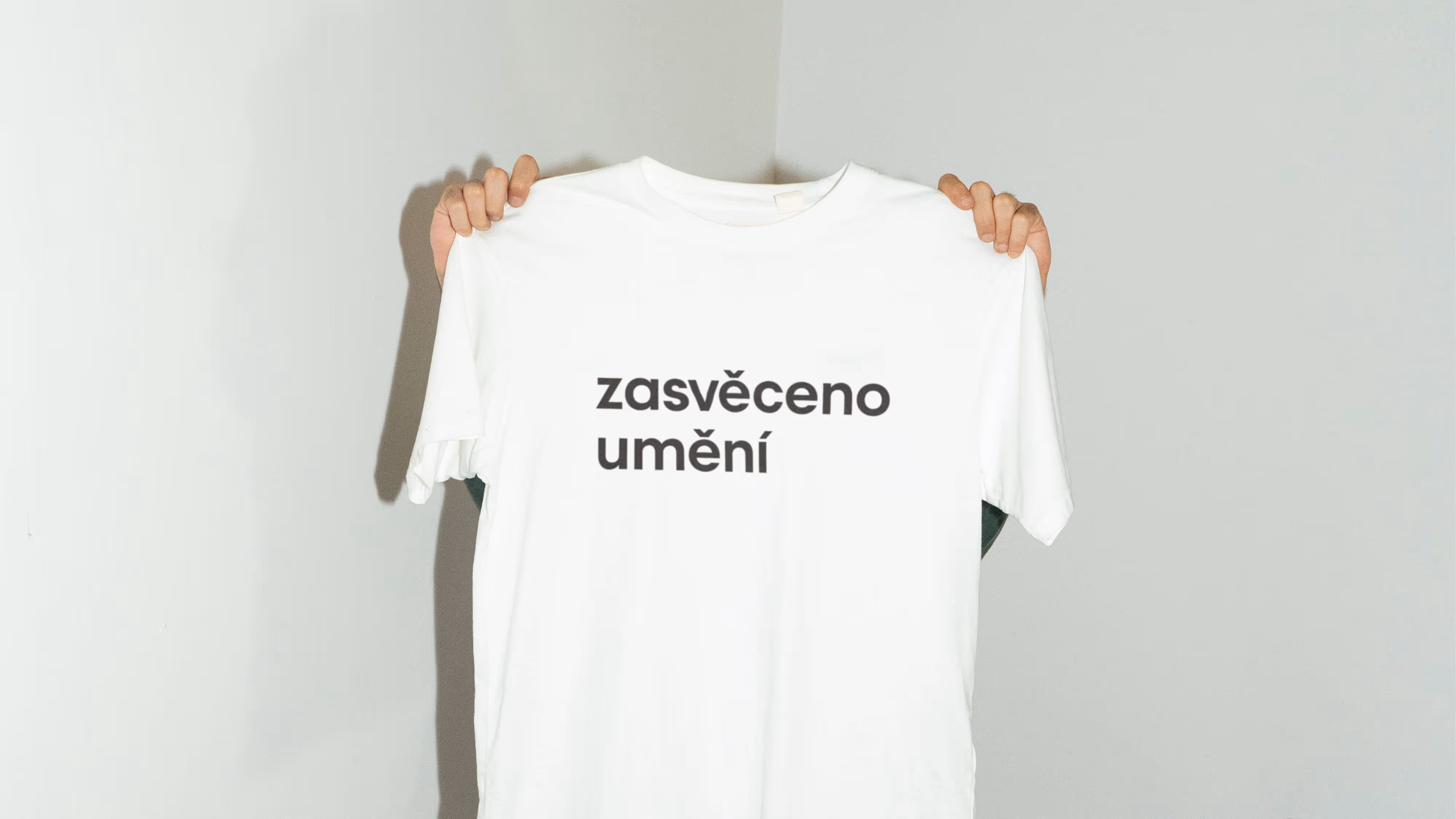 Osoba drží bílý tričko s textem „zasvěceno umění“ na jasném pozadí.