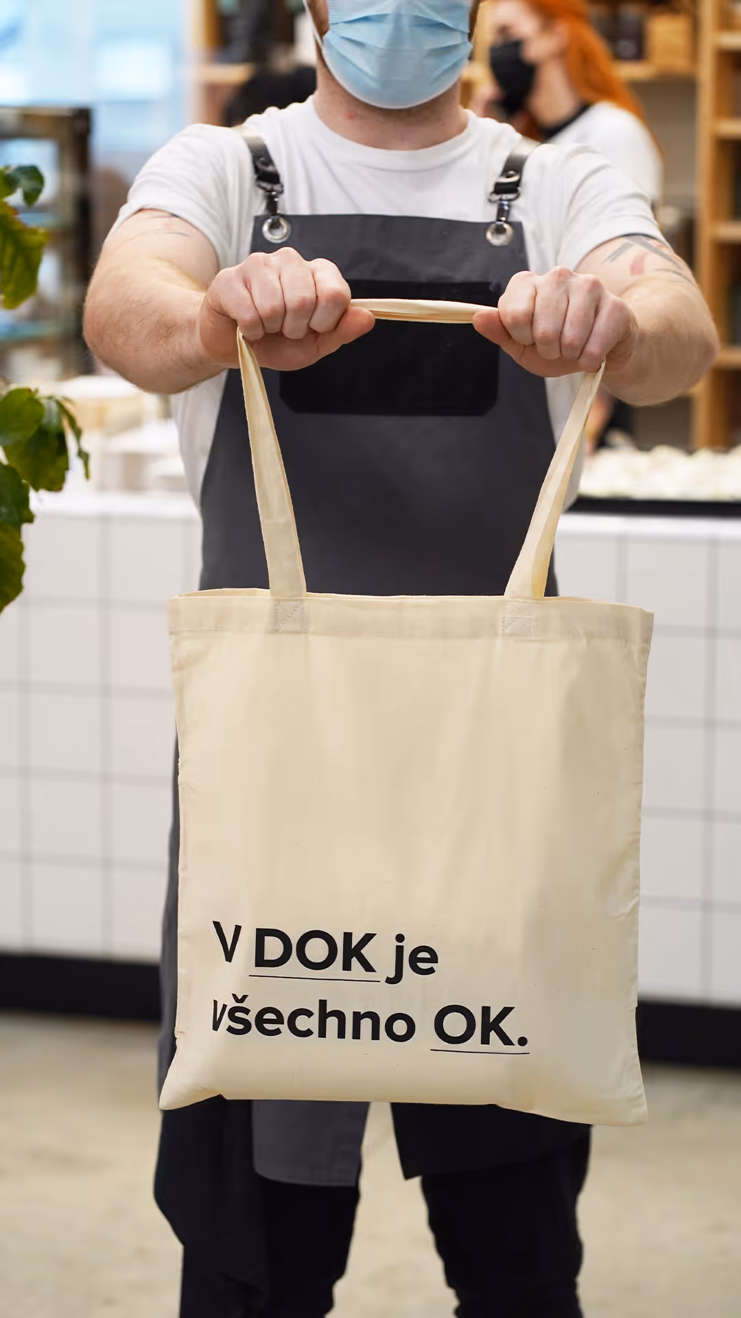 Osoba v roušce drží látkovou tašku s nápisem ‚V DOK je všechno OK.‘