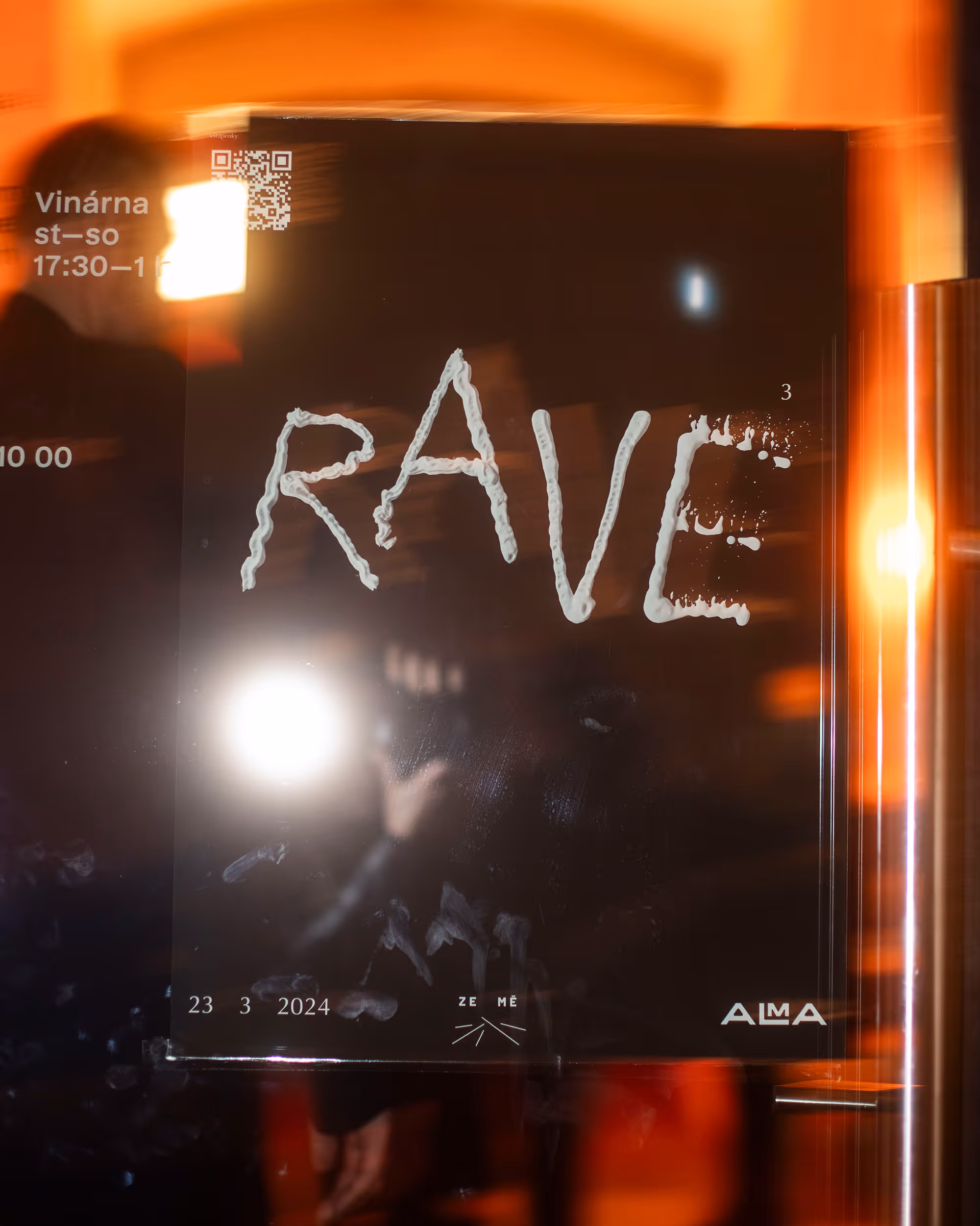 Plakát s nápisem RAVE a datem 23. 3. 2024 s oranžovým odrazem světla v pozadí.