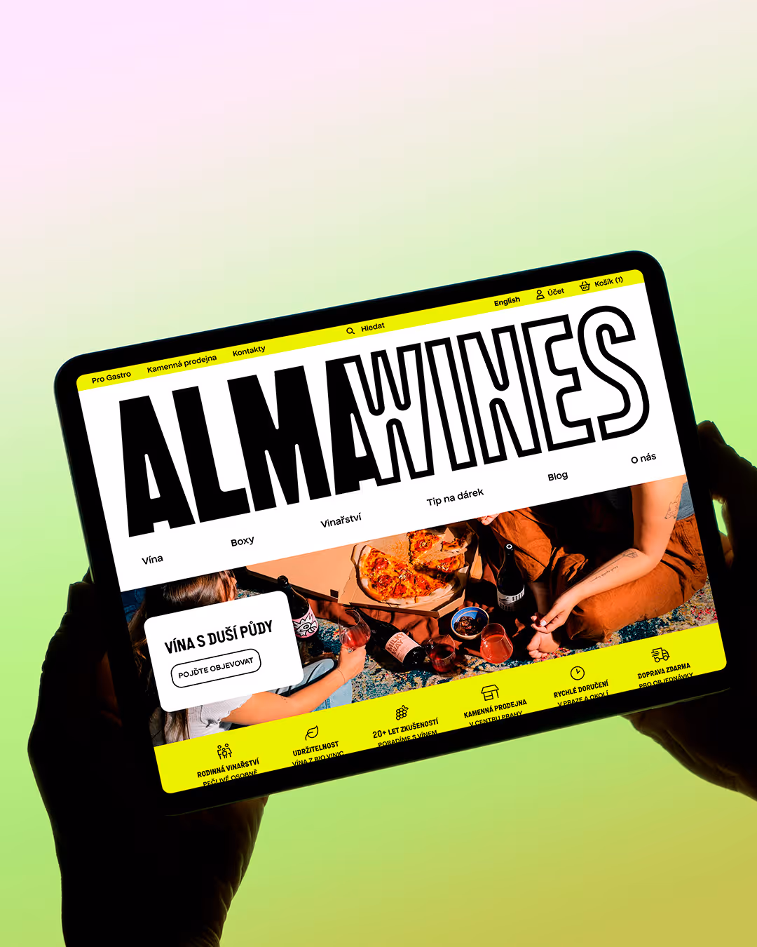 Osoba drží tablet zobrazující web ALMA WINES s vínem a pizzou na obrazovce.