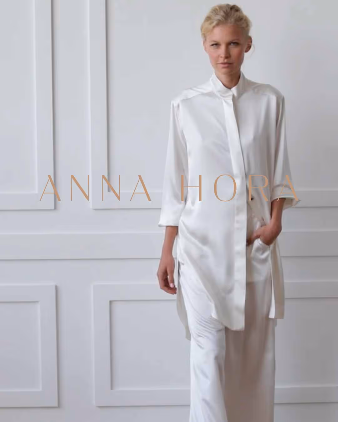 Žena v elegantním bílém saténovém outfitu stojí před zdí s dekorativními obklady.