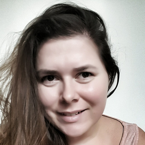Žena s dlouhými hnědými vlasy se usmívá a dívá se přímo do kamery na světlém pozadí.