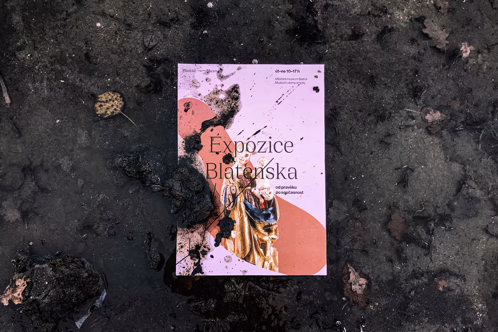Leták Expozice Blatenska ležící na blátě a mokré zemi s textem a sochou.