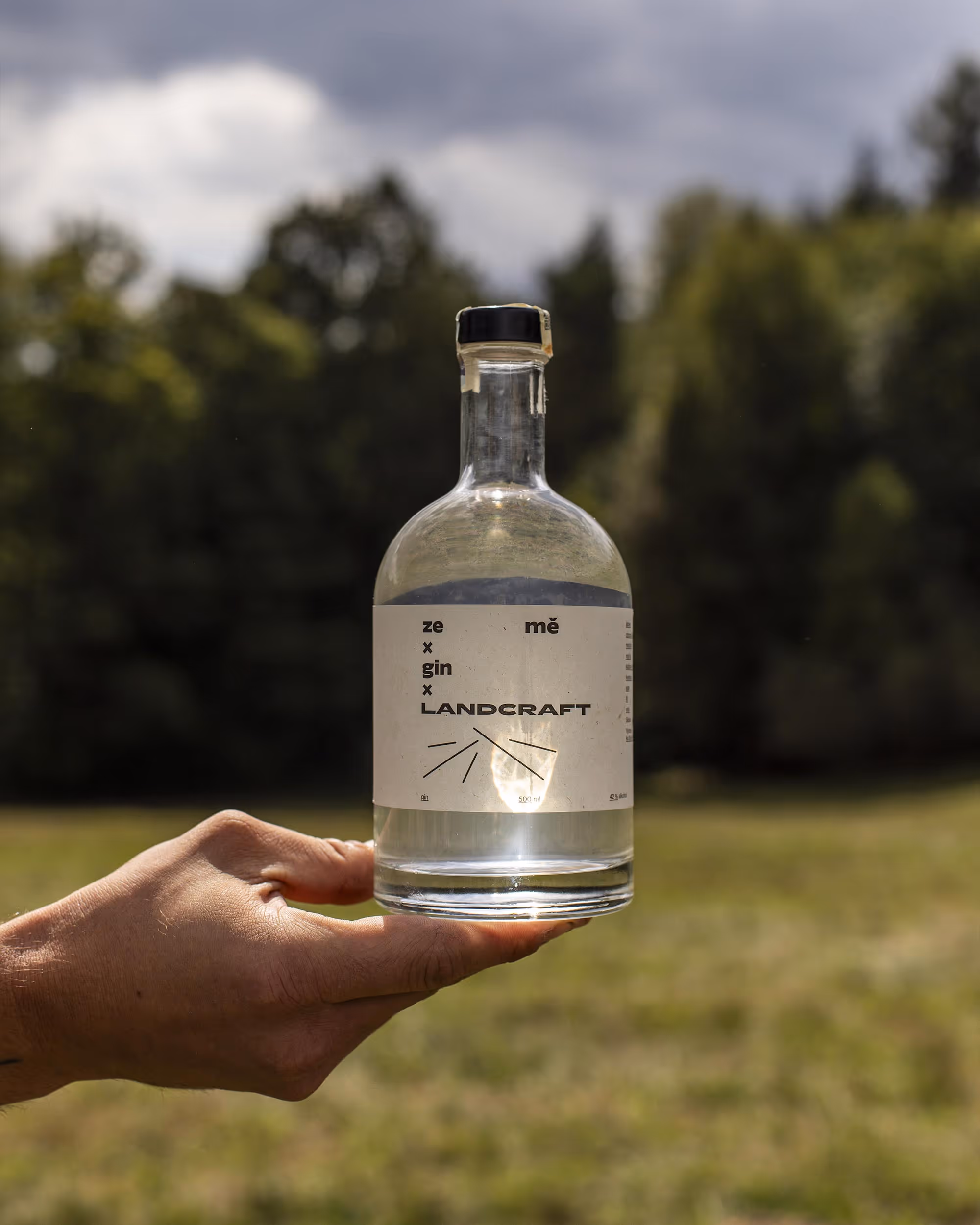 Ruka drží čistou láhev s nápisem ze x gin x LANDCRAFT na venkovním pozadí lesa.