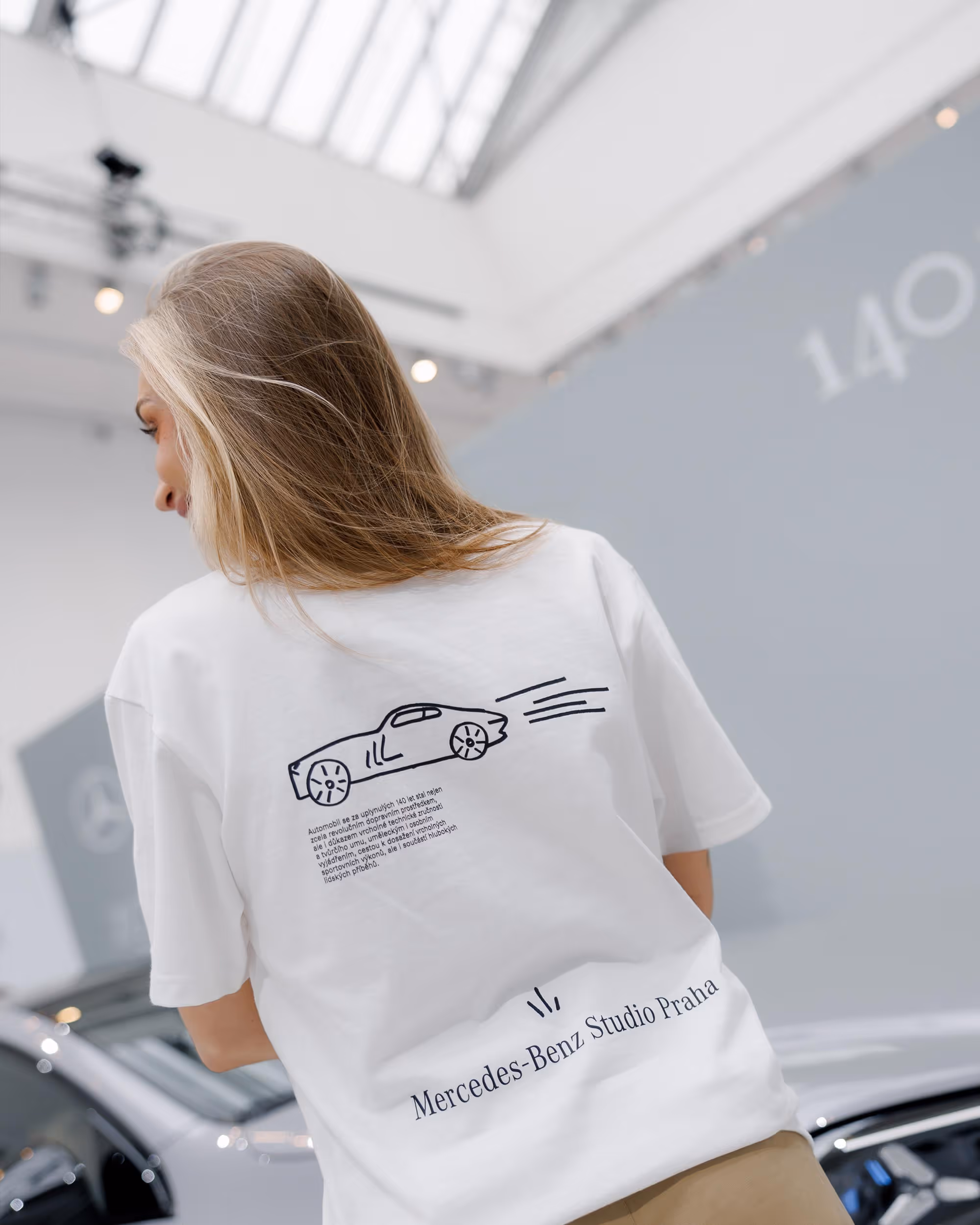 Žena zobrazená zezadu v bílem tričku s kresbou auta a nápisem Mercedes-Benz Studio Praha.