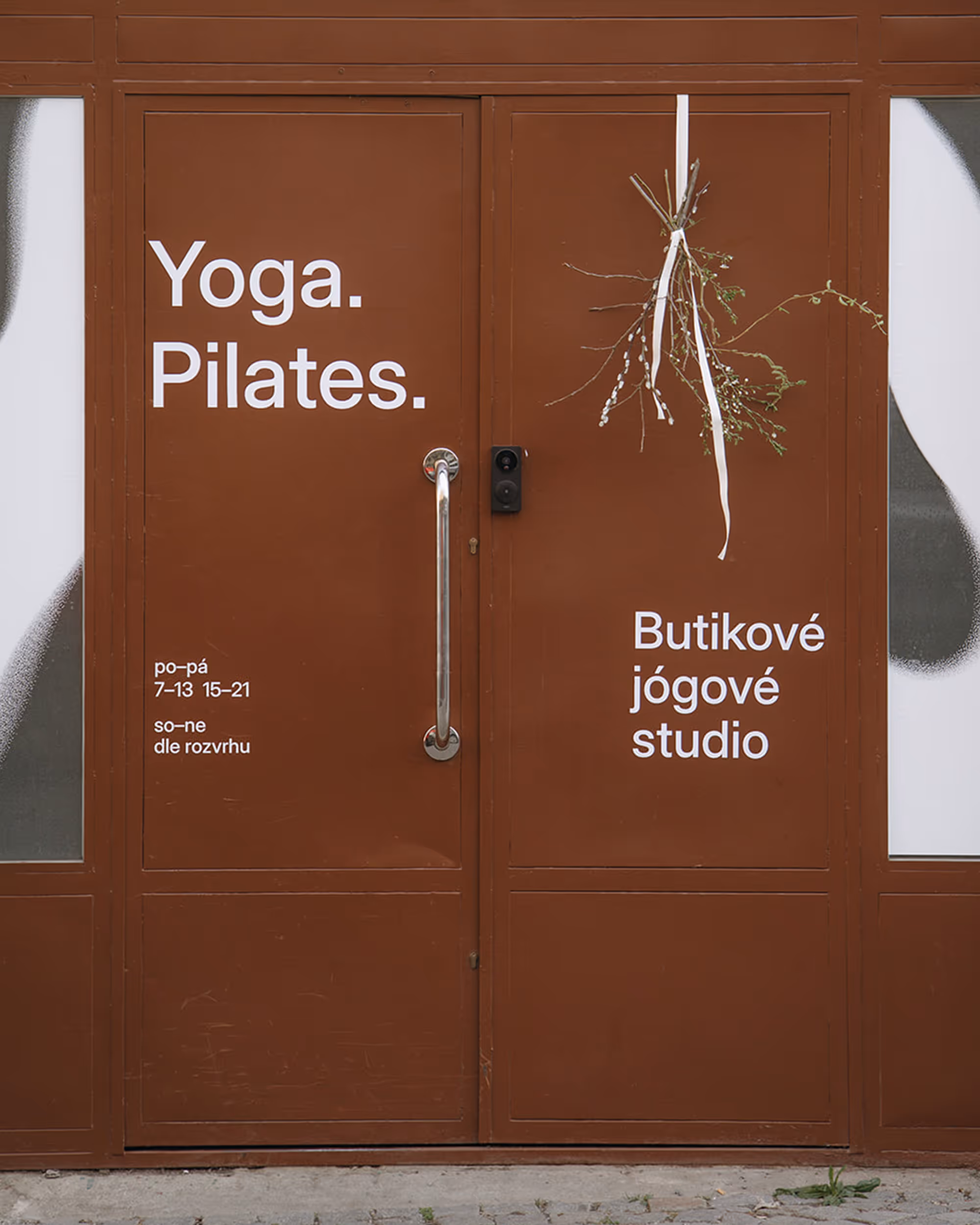 Hnědé dveře s nápisy Yoga. Pilates. a Butikové jógové studio, s větvičkami a bílou stuhou.
