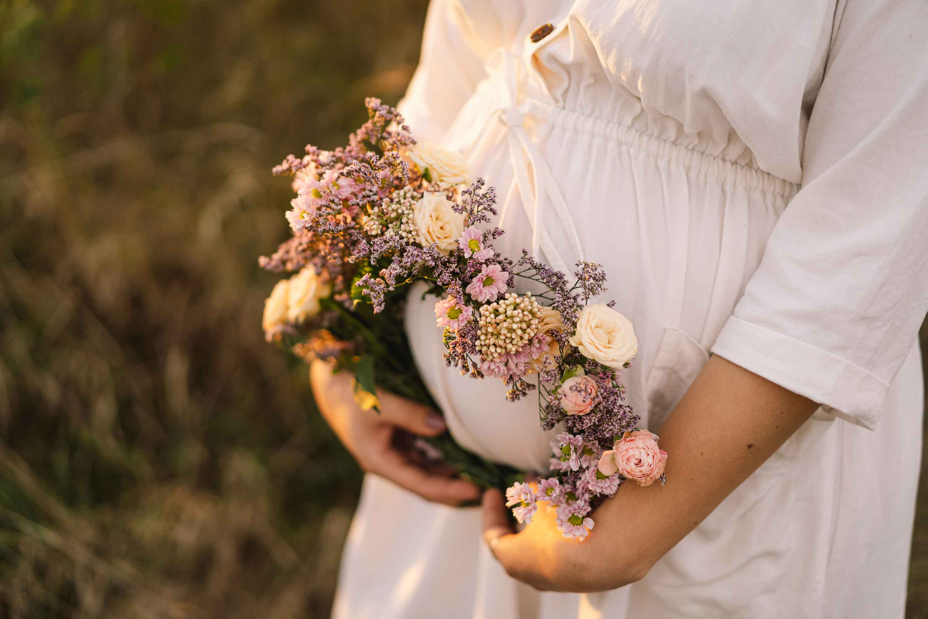 Pregnant Woman in a flowing white dress, holding a floral wreath over her baby bump.