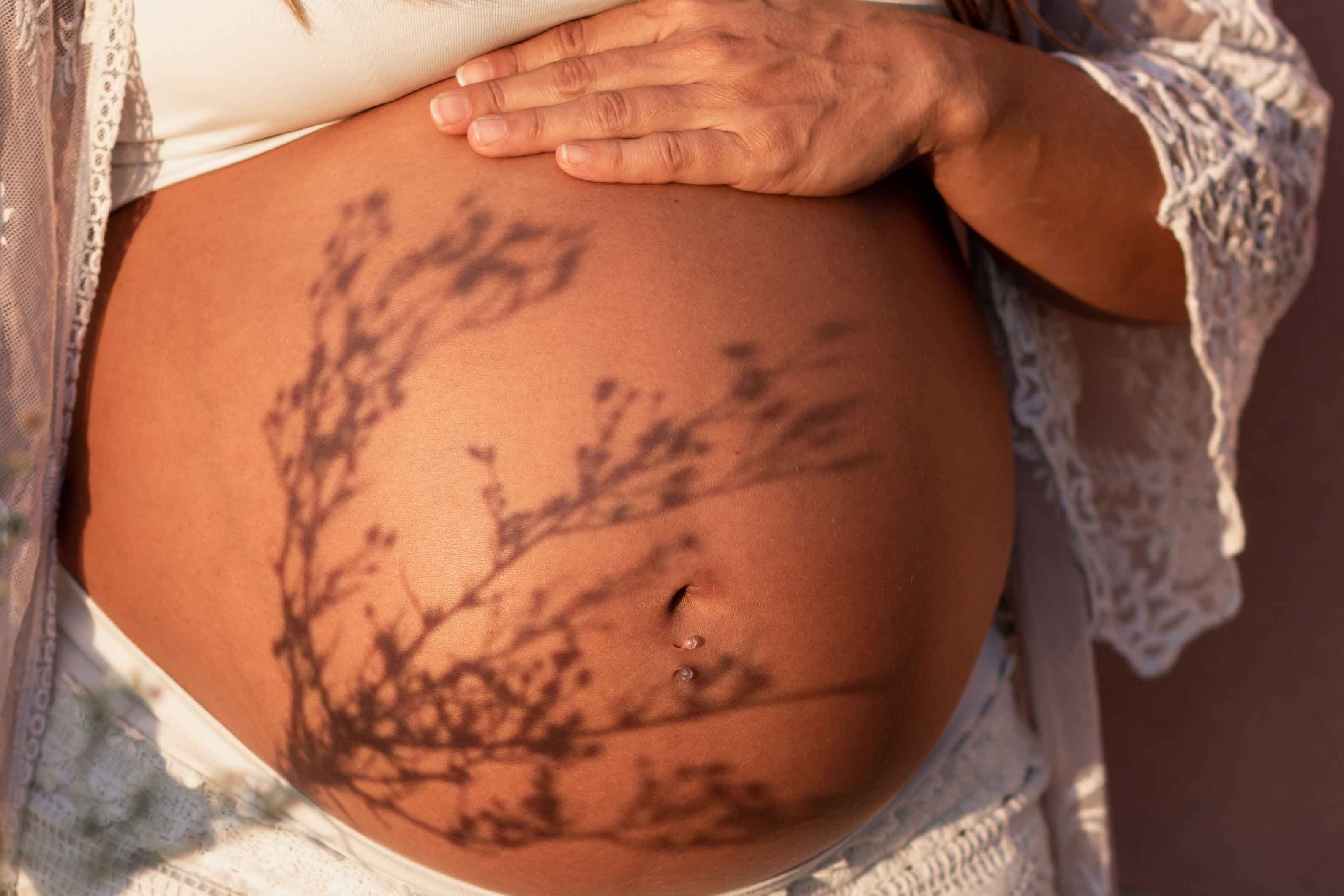 Pregnant woman showing her belly in the sunlight with a silhouette of flowers casted on her belly