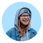 Smiling person with glasses wearing a blue hooded jacket and gray hoodie against a light blue background.
