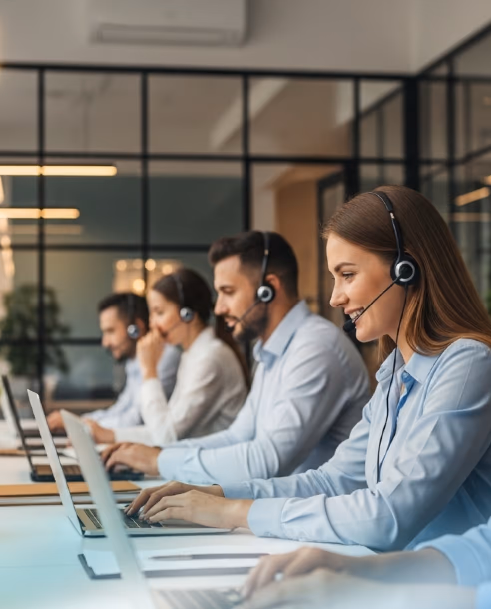 Row of customer service representatives wearing headsets and working on laptops in an office.