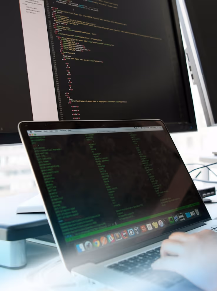 Laptop displaying code and data with hand typing, in front of a larger monitor also showing programming code.