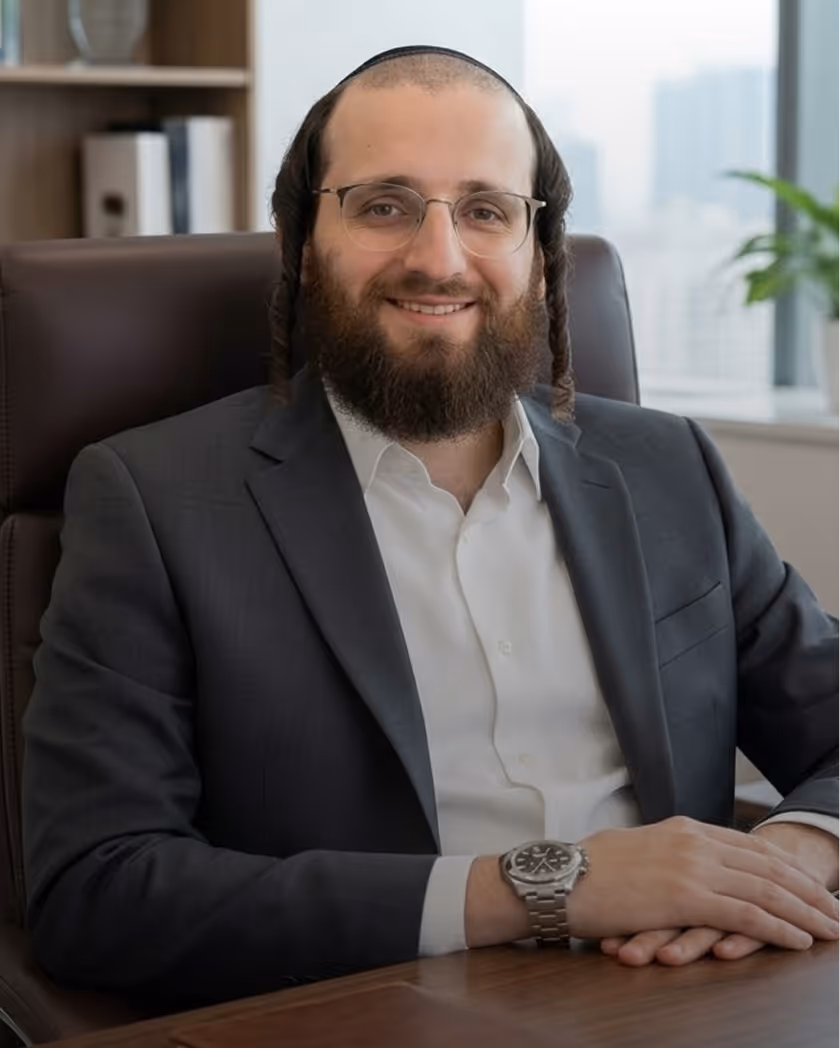 Smiling man with glasses, beard, and sidelocks wearing a dark suit and white shirt seated at an office desk.