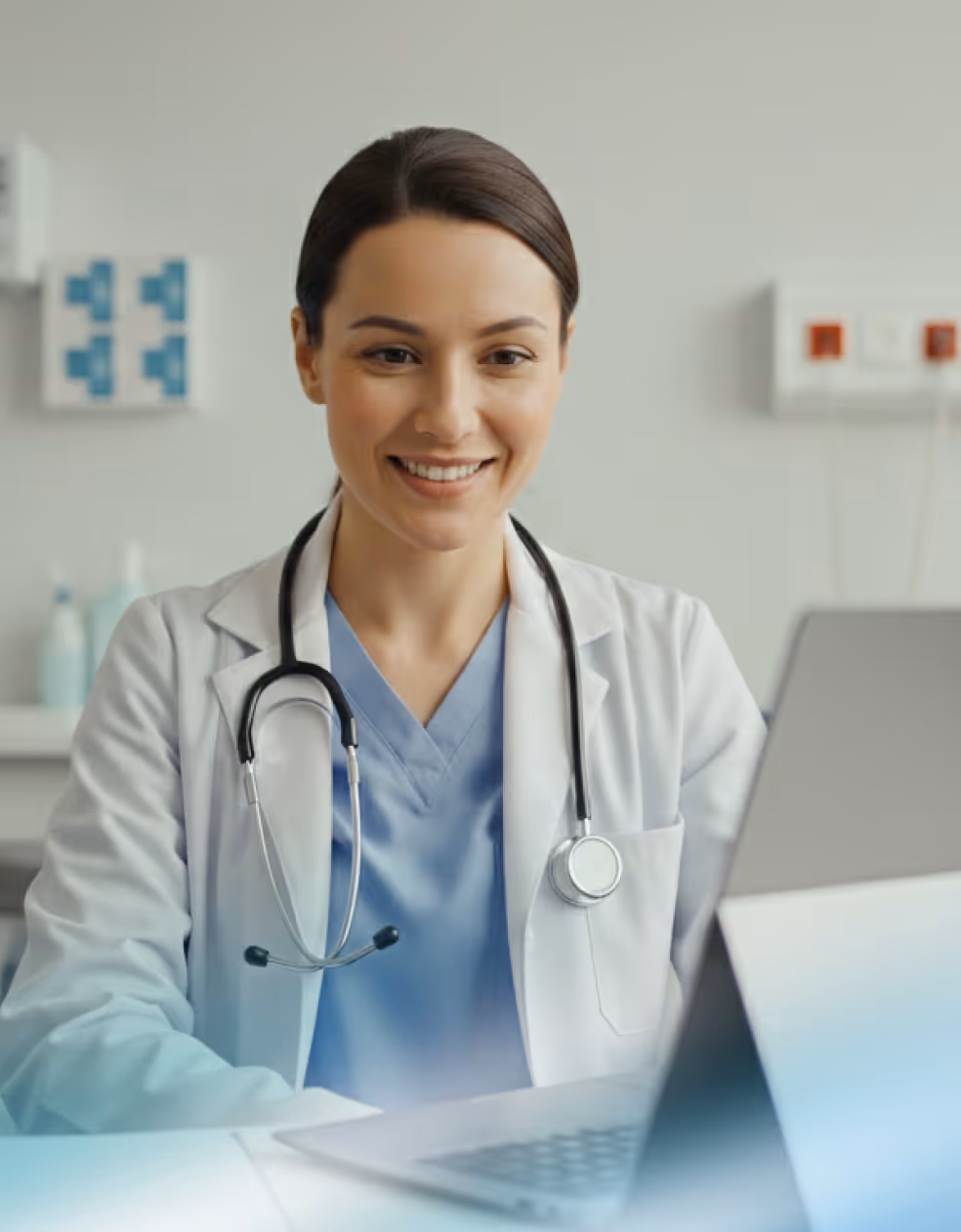 Smiling female doctor wearing a white coat and stethoscope, looking at a laptop in a medical office.