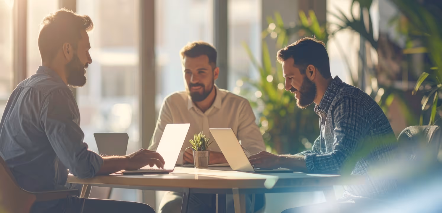 Three men with beards working on laptops and smiling at a round table in a sunlit office with plants.