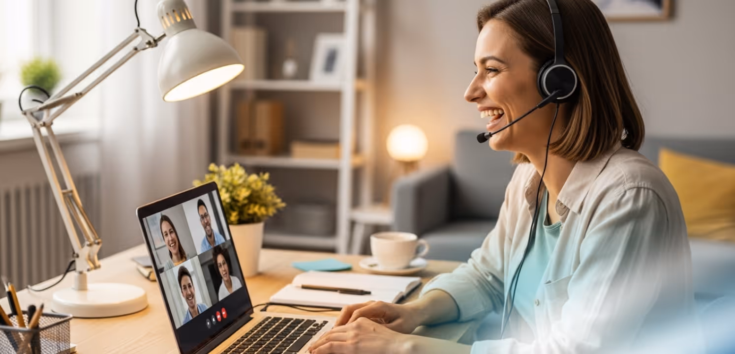 Woman wearing headset smiling and participating in a video call with four people on a laptop in a cozy home office.