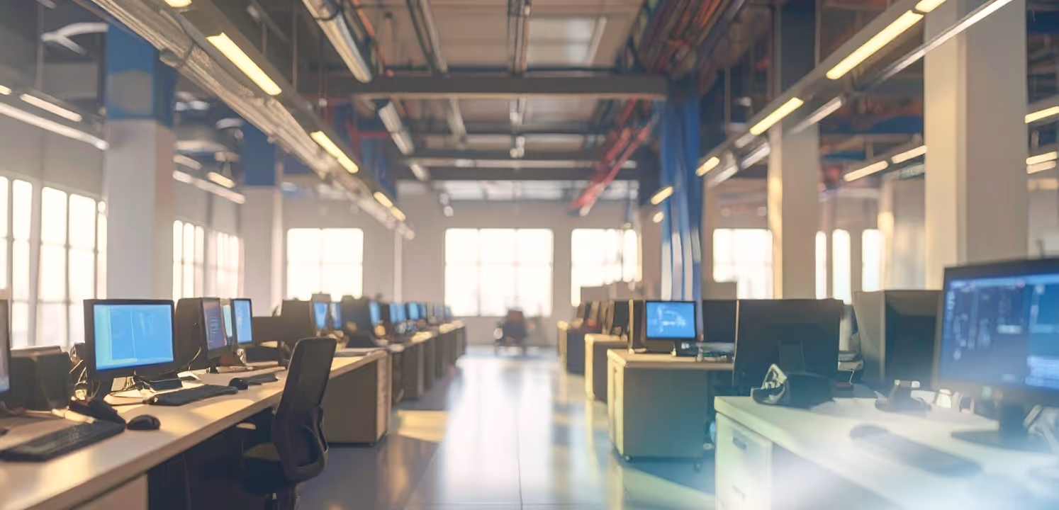 Empty modern office with rows of desks and computer monitors illuminated by natural light.