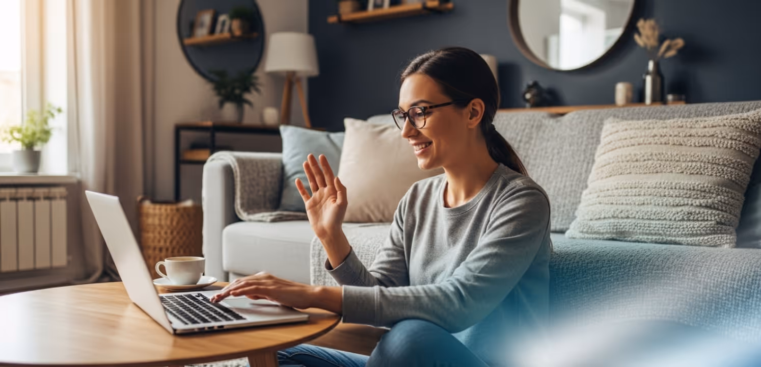 Woman sitting cross-legged on the floor in a cozy living room, waving and smiling at a laptop on a wooden coffee table.