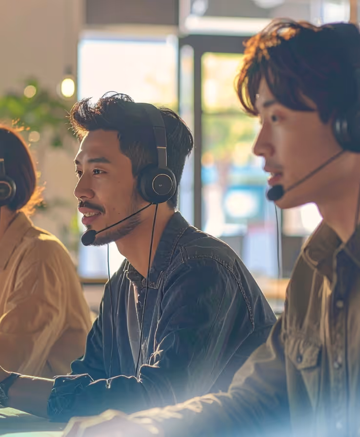 Three people wearing headsets working in a bright office, engaged in call center or customer support tasks.