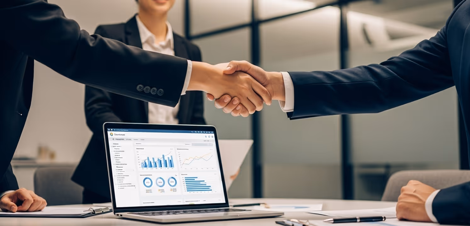 Two businesspeople shaking hands over a table with a laptop displaying financial charts and graphs.