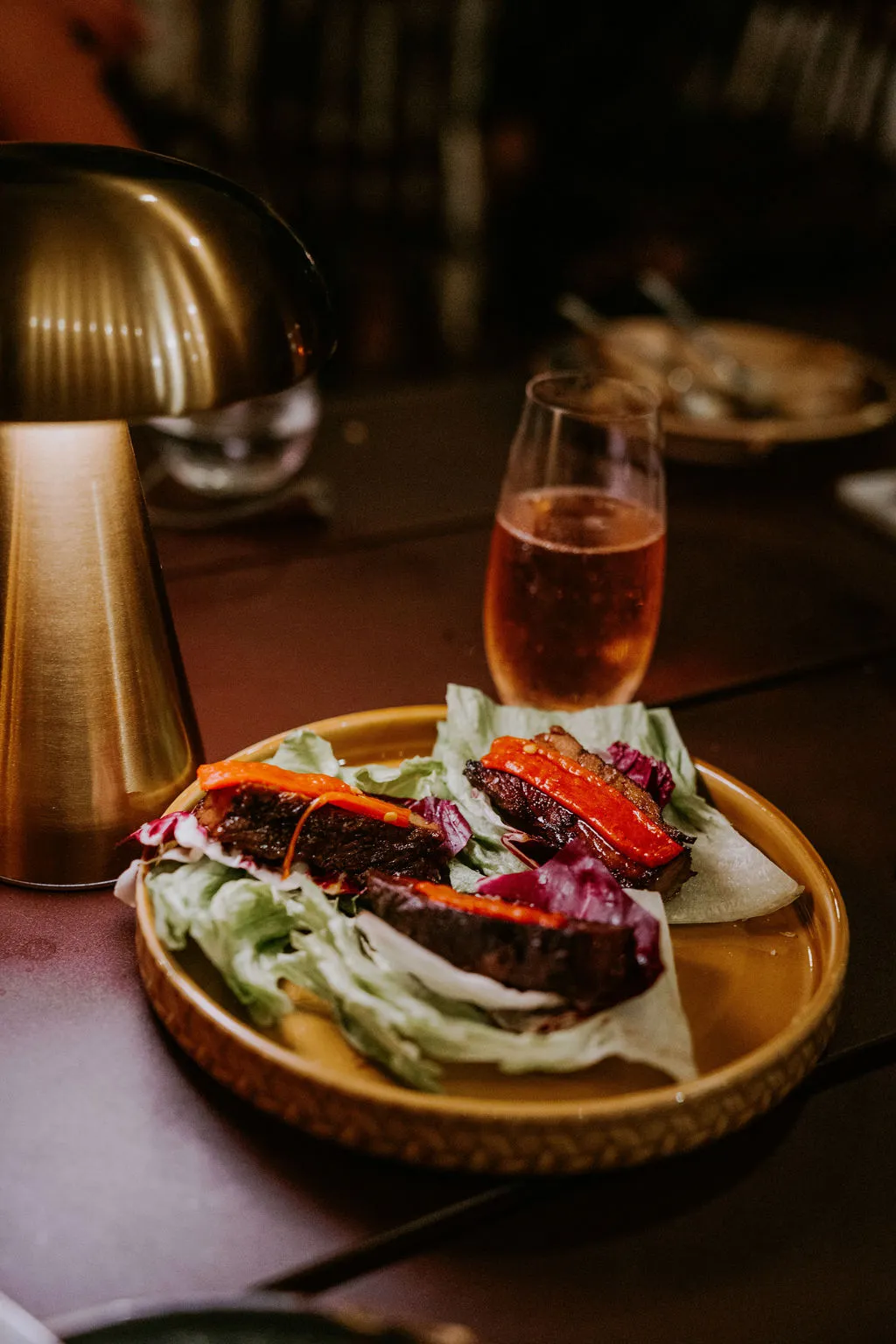Plate with three lettuce wraps filled with grilled meat and red pepper slices, next to a golden lamp and a glass of rosé wine on a dark table.